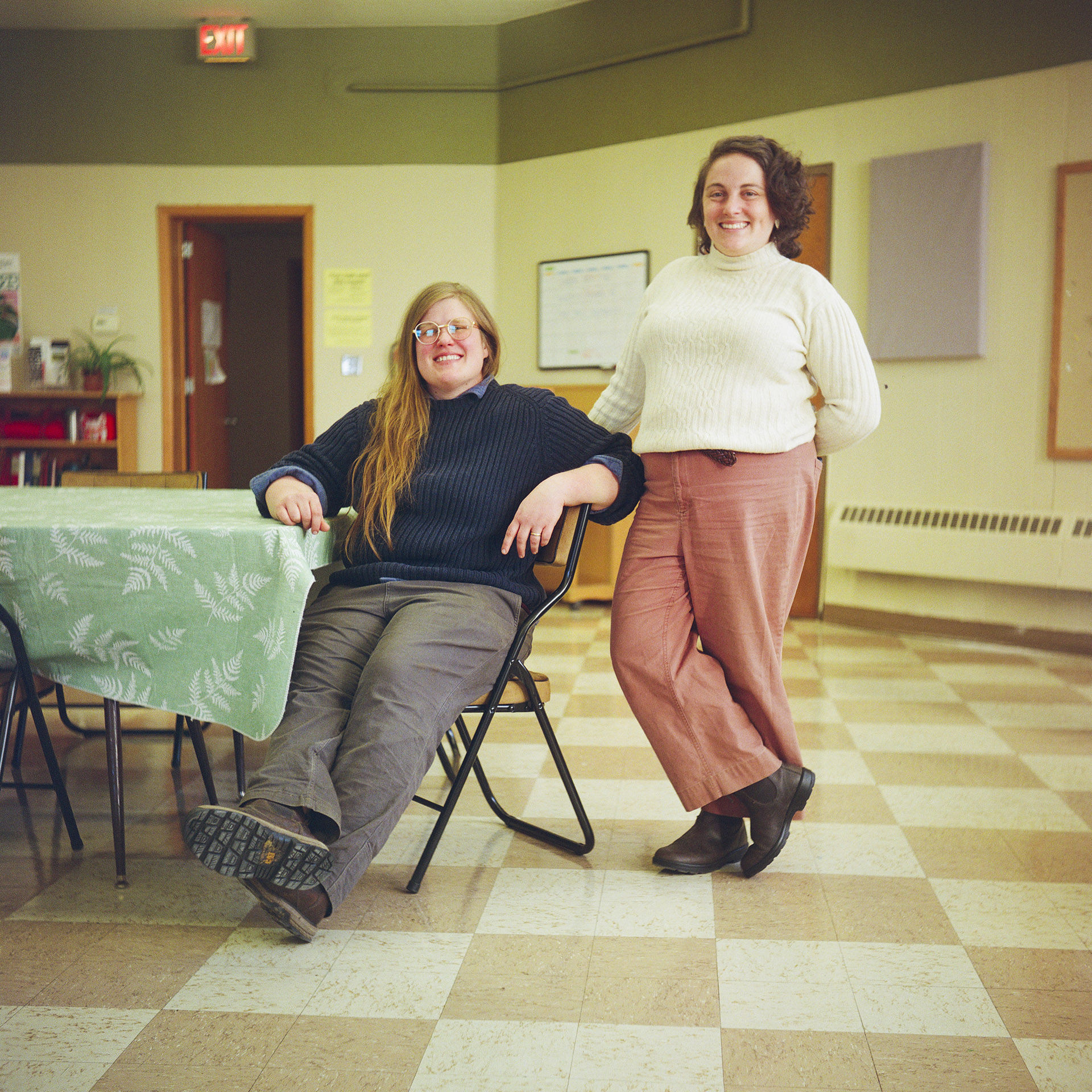 Rebecca Krausert Sykalski sits in a folding metal chair next to a table covered by a tablecloth and Rachel Peller stands next to her in a room with vinyl panel flooring, and with an open door next to a bookshelf on a rear wall with an illuminated "EXIT" sign mounted to the ceiling.