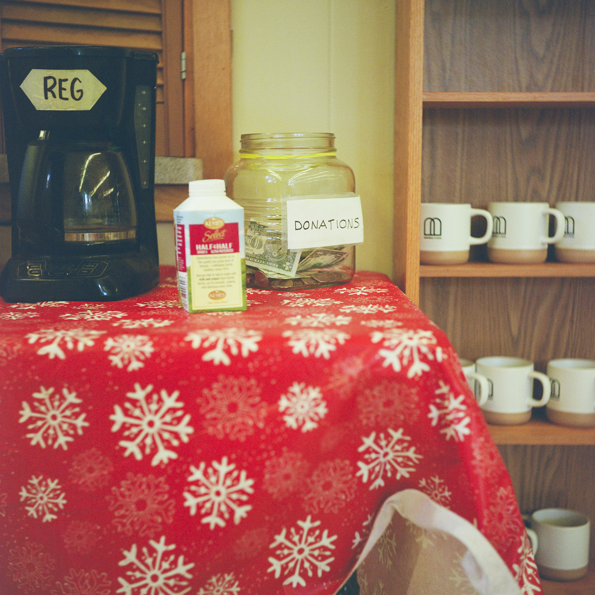A plastic jar with the word "Donations" written on a piece of paper taped to its side sits next to a carton of half-and-half and a drip coffee maker with the word "Reg" written on tape affixed to its side on top of a snowflake-themed tablecloth, with shelf with ceramic mugs on multiple levels in the background.