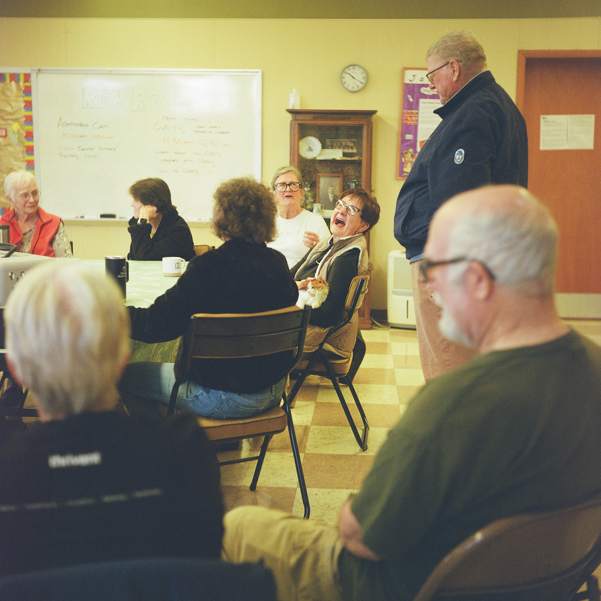 Jill Roethe sits in a padded metal folding chair, with other people seated on other side of her and in the foreground, and with one person standing behind her, in a room with a large whiteboard mounted to a wall next to a display cabinet, clock, sign and closed door.
