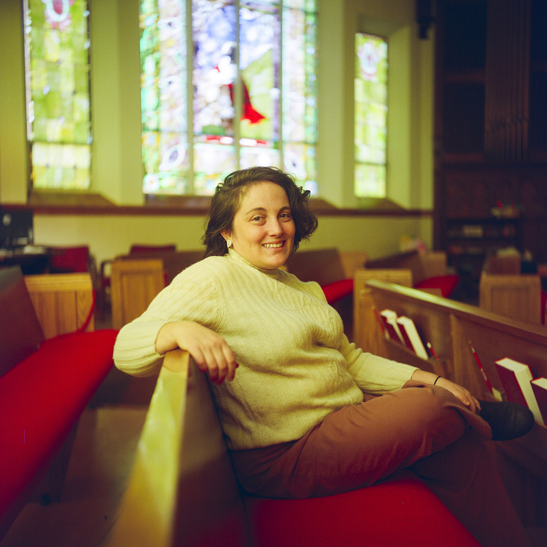 Rachel Peller sits on a padded wood pew, with other pews in front of and behind her and in the background in a another row beneath an out-of-focus stained glass window.