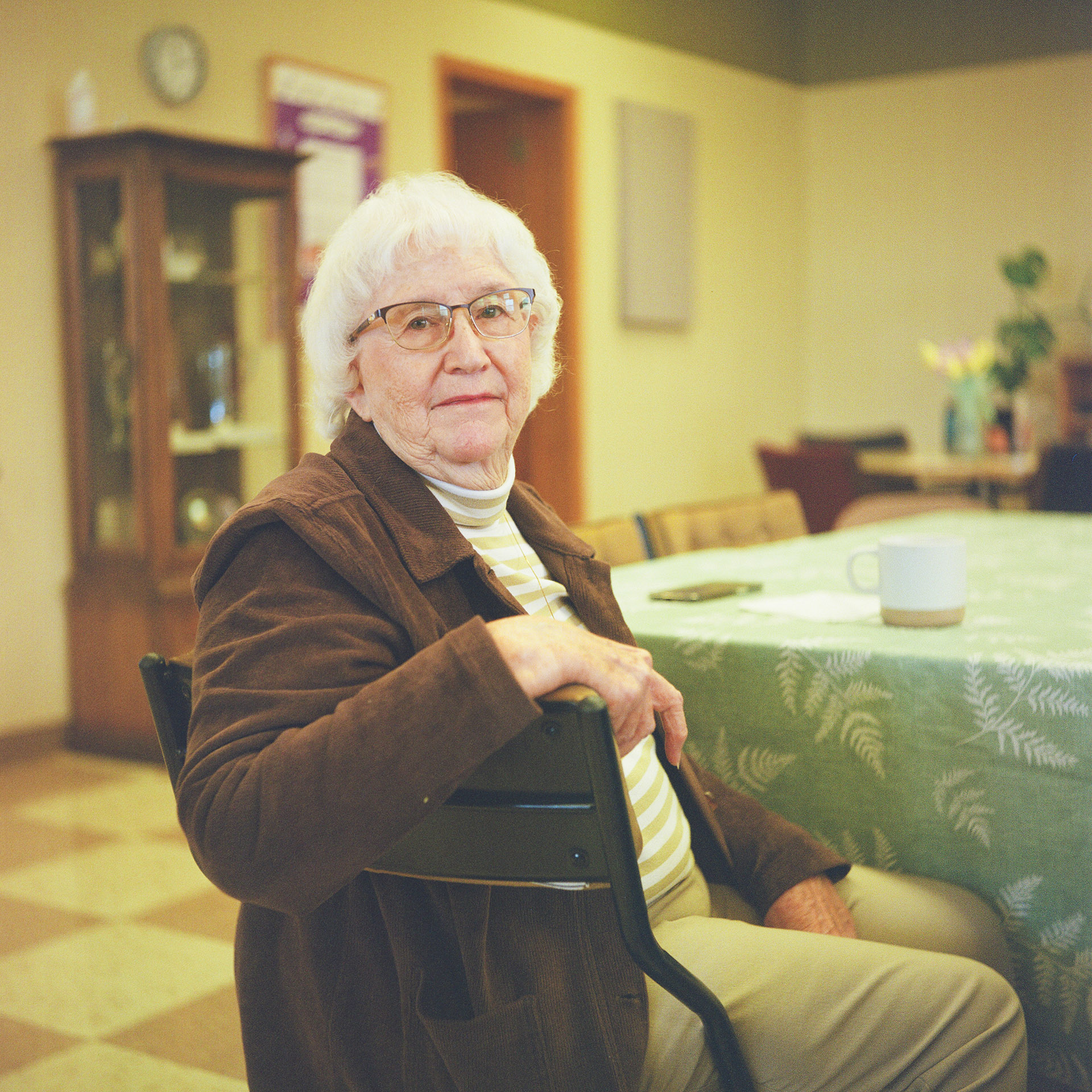 Jan Helmich sits on a padded folding metal chair next to a table covered with a tablecloth and a ceramic mug on its surface, with an out-of-focus display cabinet, clock, sign, door and table with floral displays on its surface in the background.