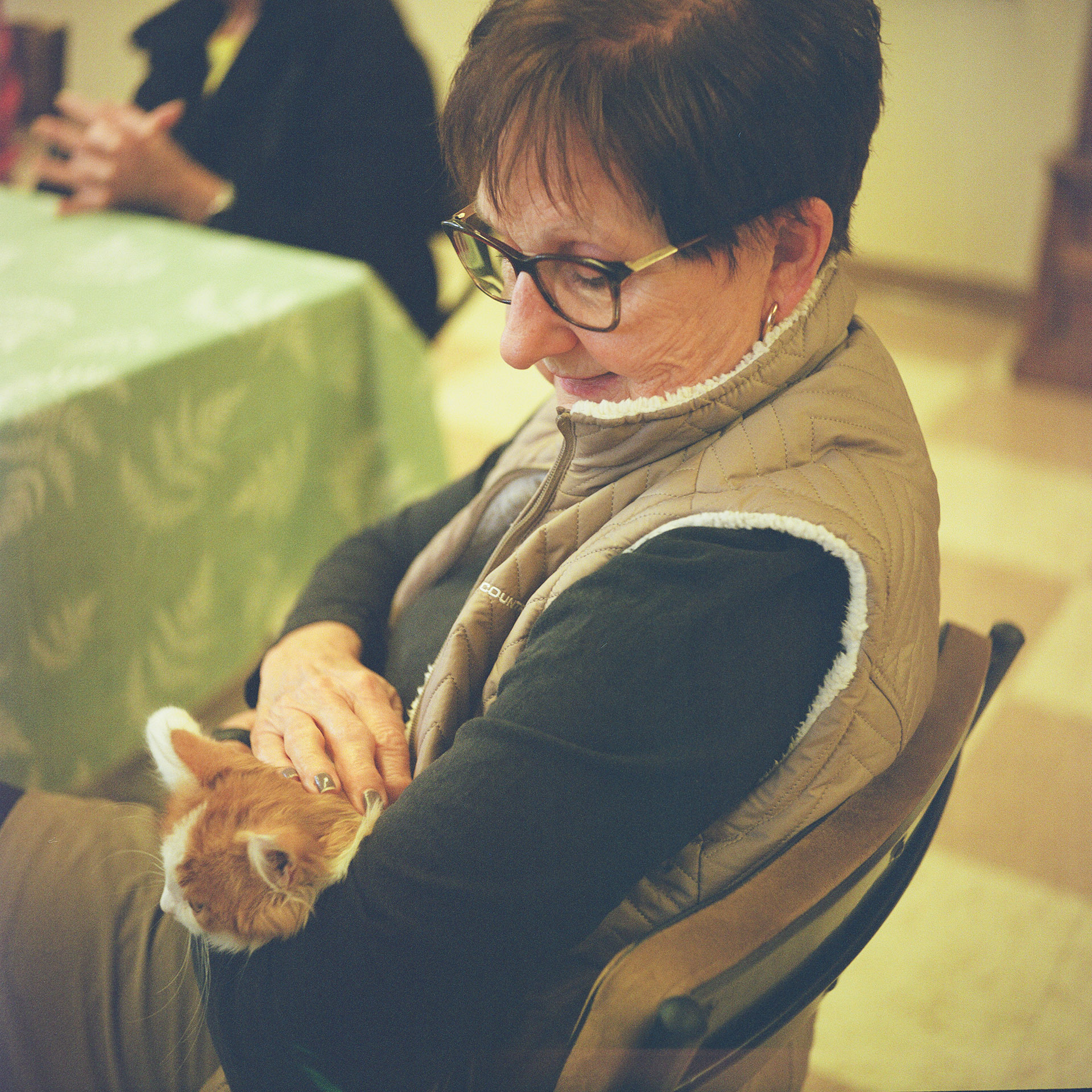 Jill Roethe holds a cat in her lap while sitting in a padded folding metal chair next to a table covered by a tablecloth, with another person sitting with folded hands on its surface.