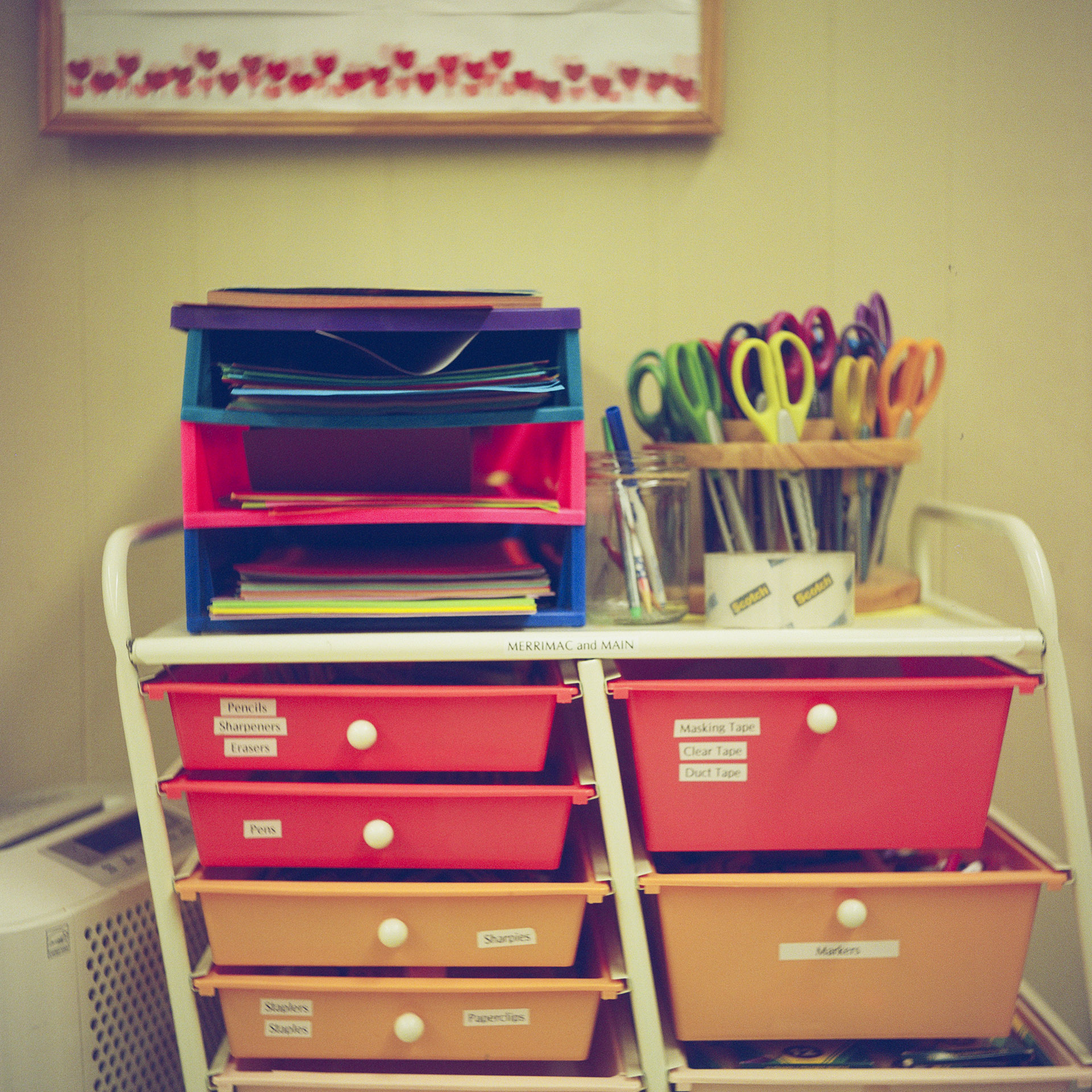 A stack of three plastic paper filing trays sits next to a glass with several markers, a scissors storage rack and a roll of tape on the top storage rack with multiple plastic bin drawers with labels in front of a wall, with an out-of-focus image mounted above.