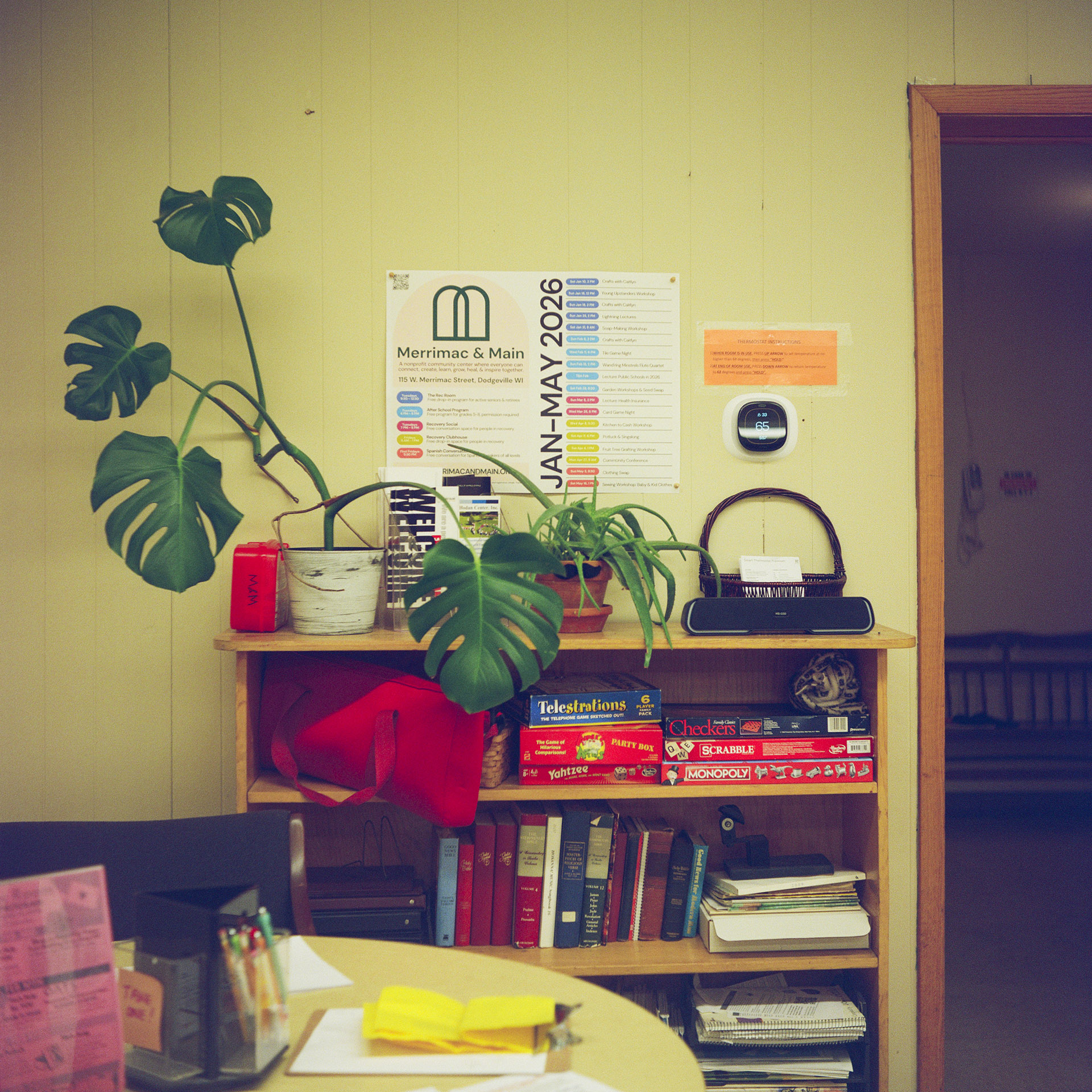 A shelf with books and games placed on different levels and with artificial plants and a wall-mounted calendar stands next to a wall next to an open door, with a table with different items on its surface in the foreground.