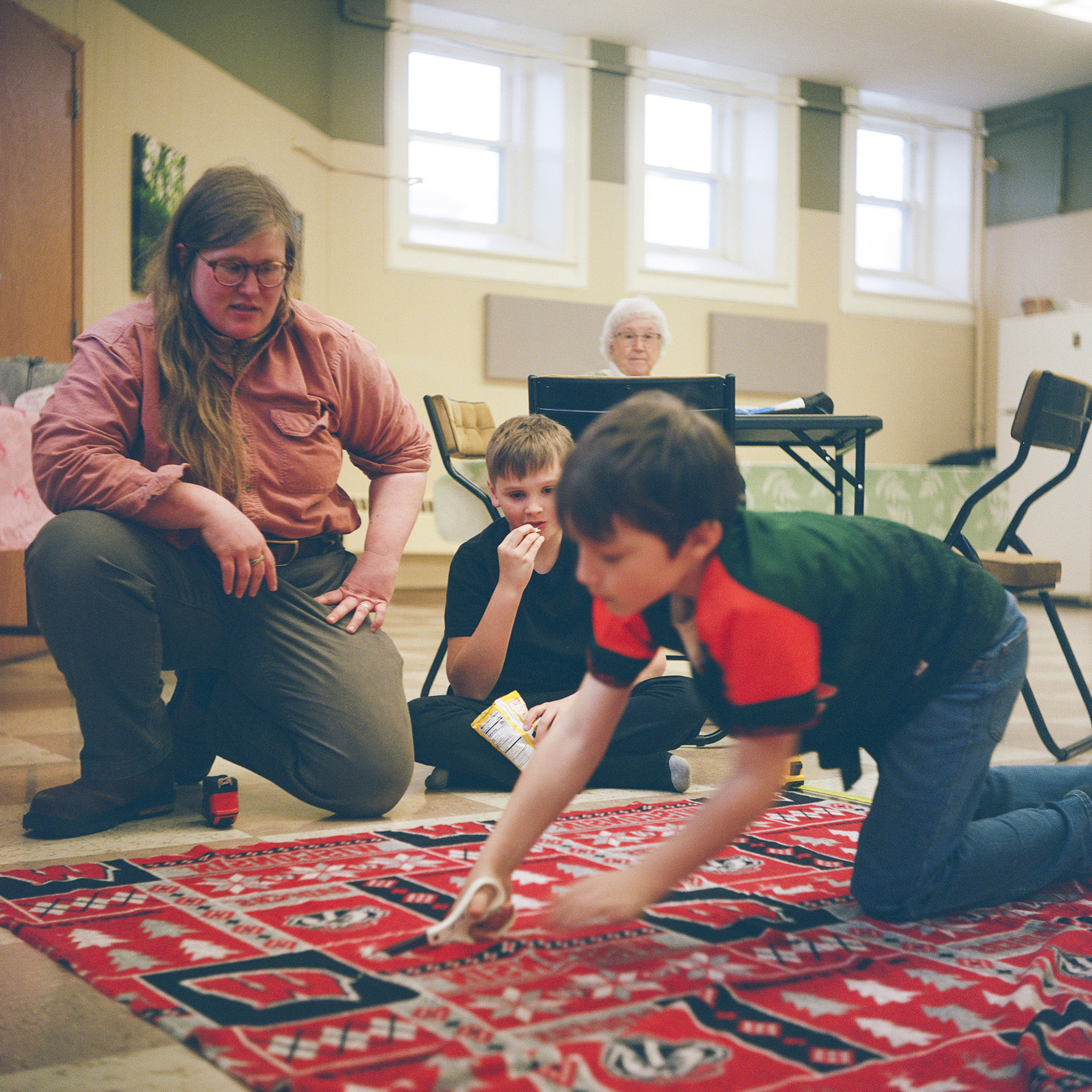 Rebecca Krausert Sykalski crouches on one kneel next to one child sitting cross-legged on the floor and with another child cutting a bolt of fabric on the surface of a floor, with another person seated at a folding table in the background, in a room with a closed door and three windows placed near the ceiling.