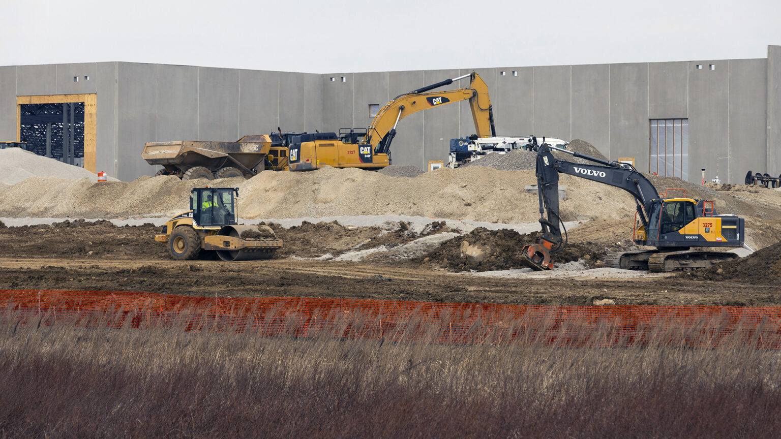 Multiple construction vehicles work among piles of gravel and dirt in front of a concrete wall, with a woven barrier fence and prairie grasses in the foreground.