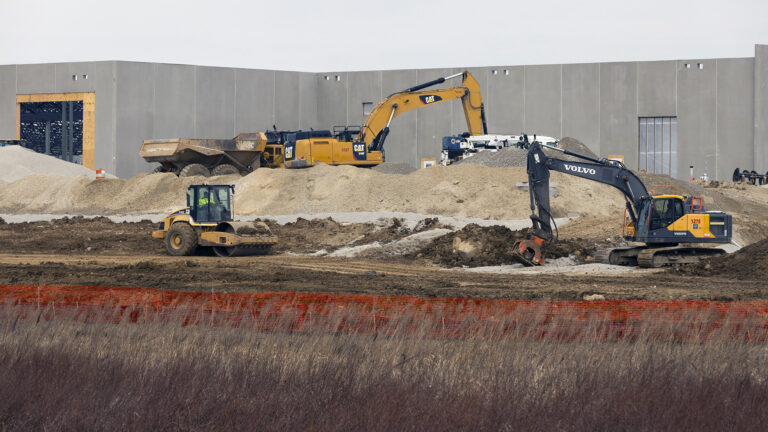 Multiple construction vehicles work among piles of gravel and dirt in front of a concrete wall, with a woven barrier fence and prairie grasses in the foreground.