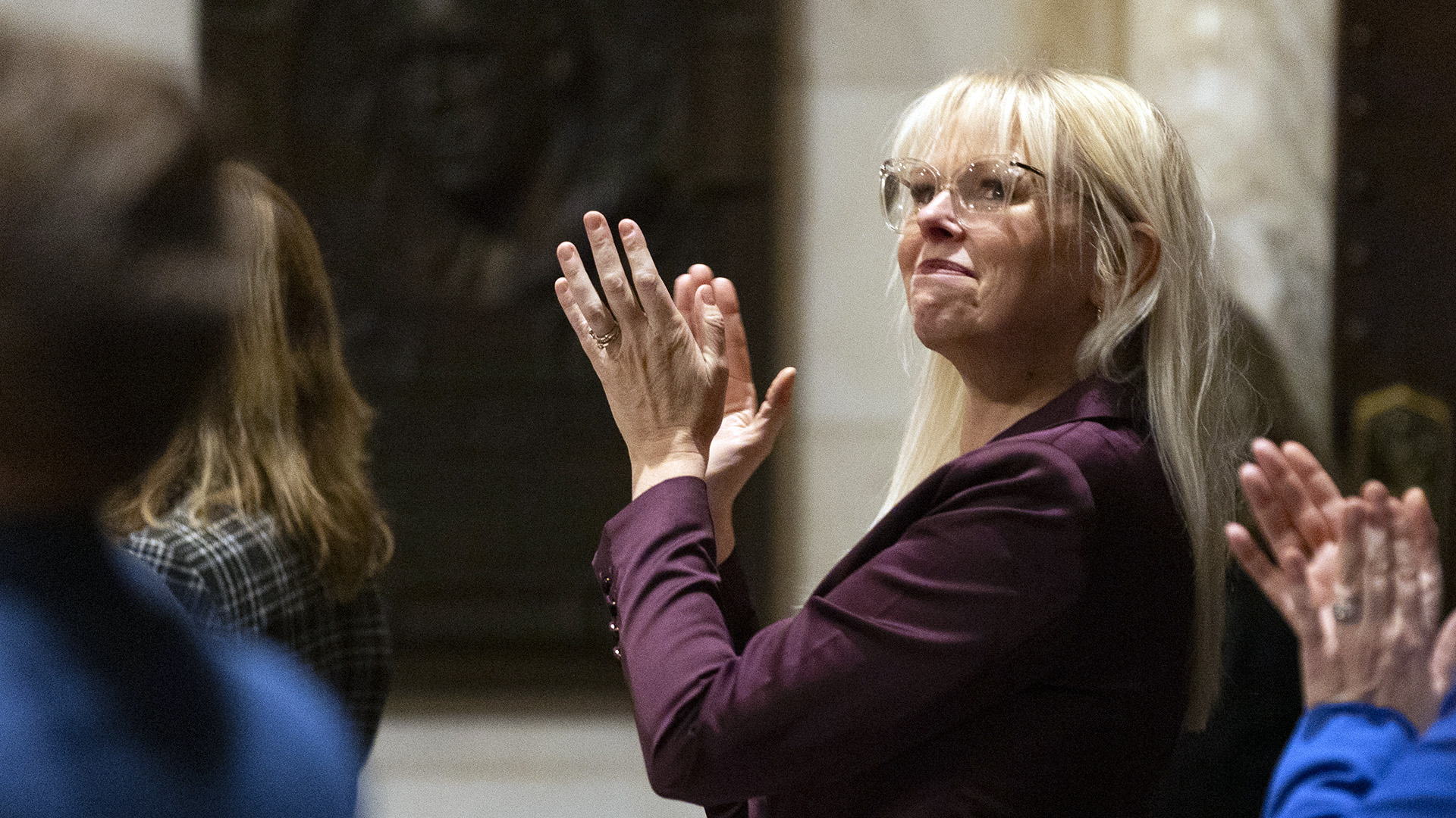 Sarah Keyeski applauds while standing, with other people standing around her, in a room with an out-of-focus bronze plaque mounted on a wall in the background.