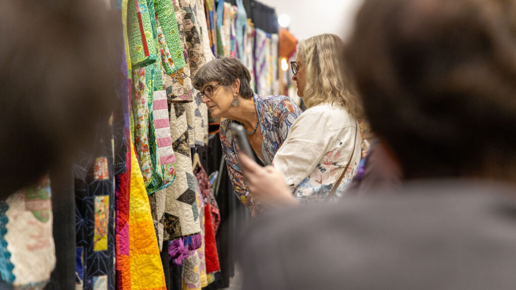 Through the blur of a crowd, two women examine quilted garments hanging in a gallery.