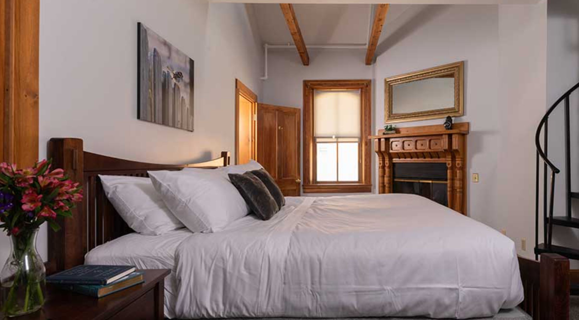 Image from Hotel Galena for featured item in Auction 2026 blog.Alt text: A cozy bedroom at Hotel Galena with a wooden bed, white linens, and decorative pillows. A fireplace, wooden beams, and a spiral staircase add rustic charm.