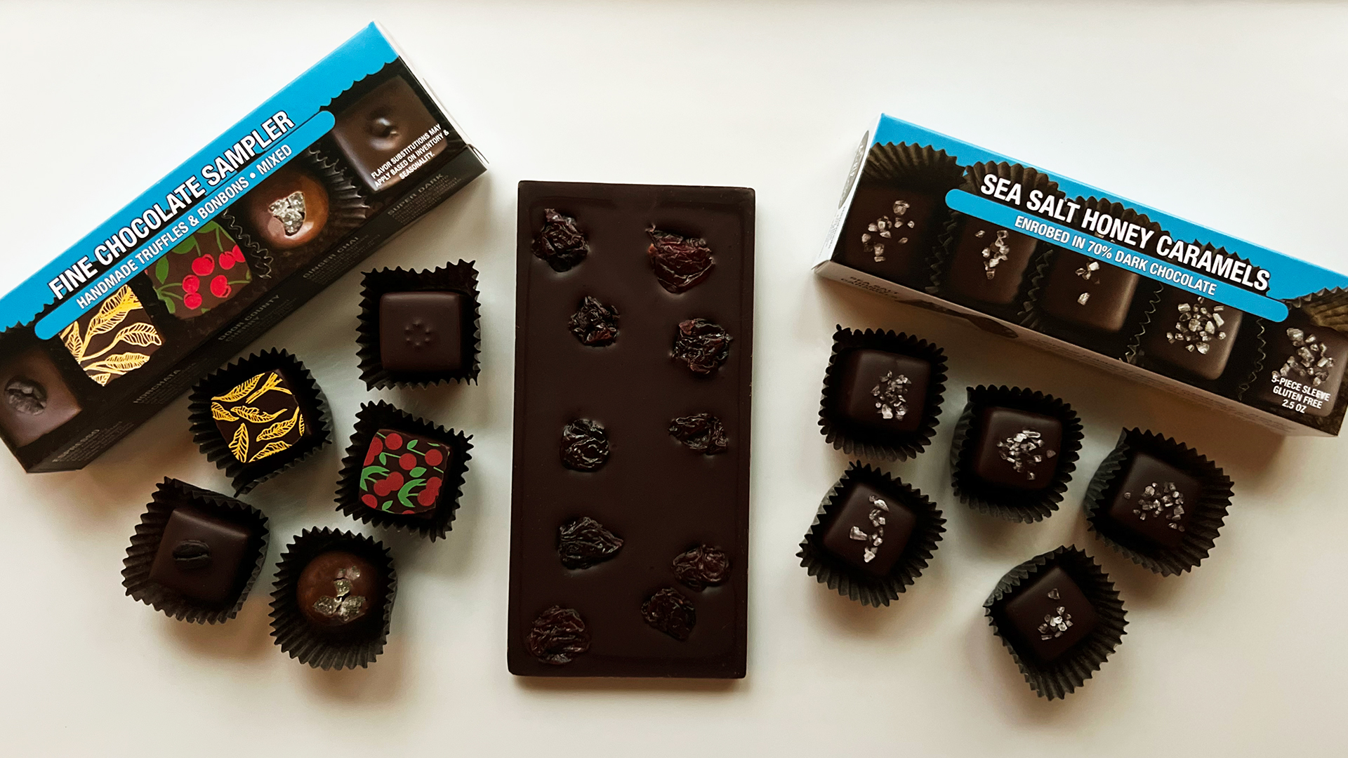 Assorted chocolates from Driftless Chocolates are displayed with two sampler boxes. A chocolate bar topped with dried cherries is centered, surrounded by individual chocolates.