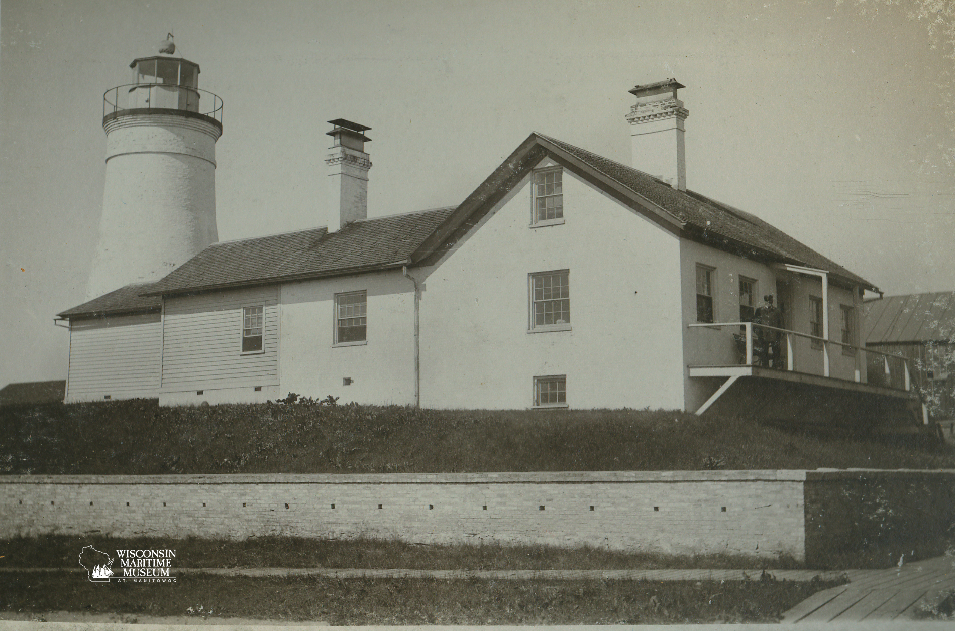 Front view of the first Manitowoc lighthouse with an attached house, featuring a tall cylindrical tower and two chimneys. The scene is calm, conveying a vintage feel. Built in 1839.