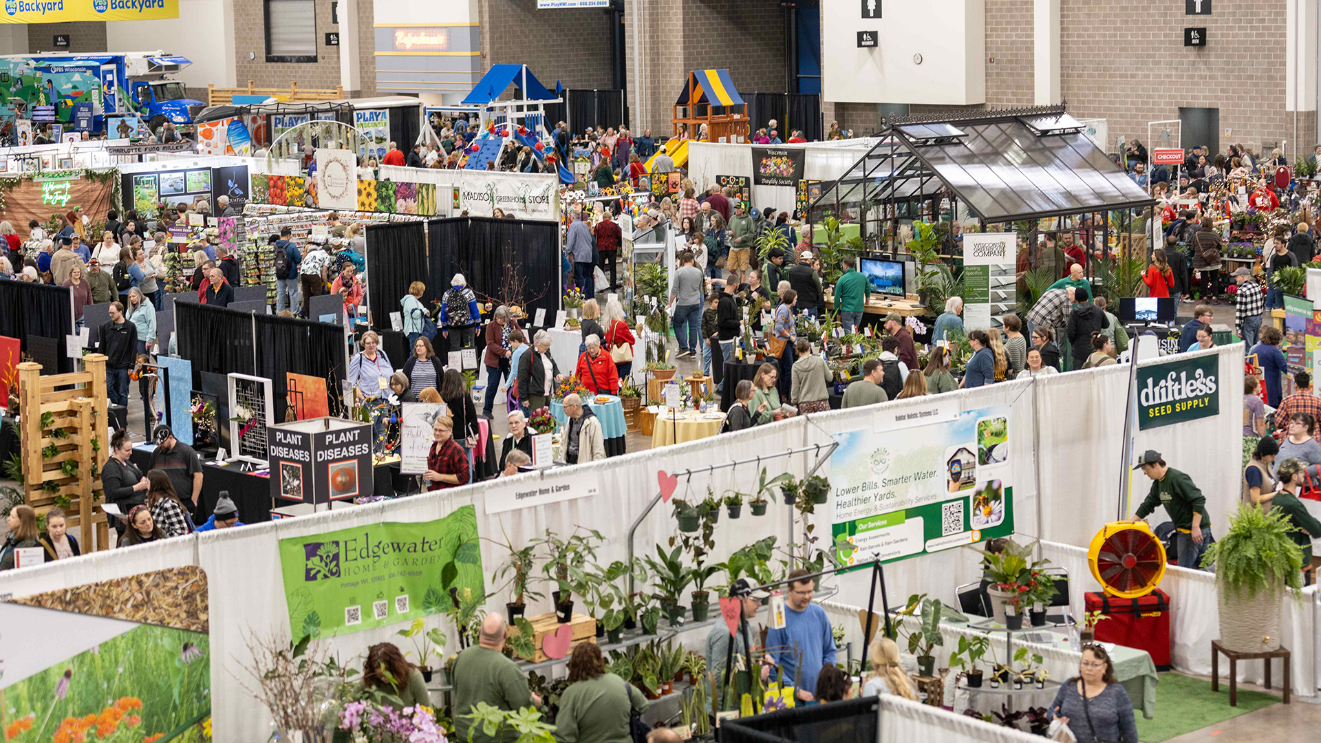 Aerial view of a crowded indoor garden and home expo, with dozens of vendor booths displaying plants, flowers, and gardening products. Visible exhibitors include Edgewater Home & Garden, Driftless Seed Supply, Madison Greenhouse Store, and a home energy sustainability services booth. Attendees browse the packed convention hall floor.