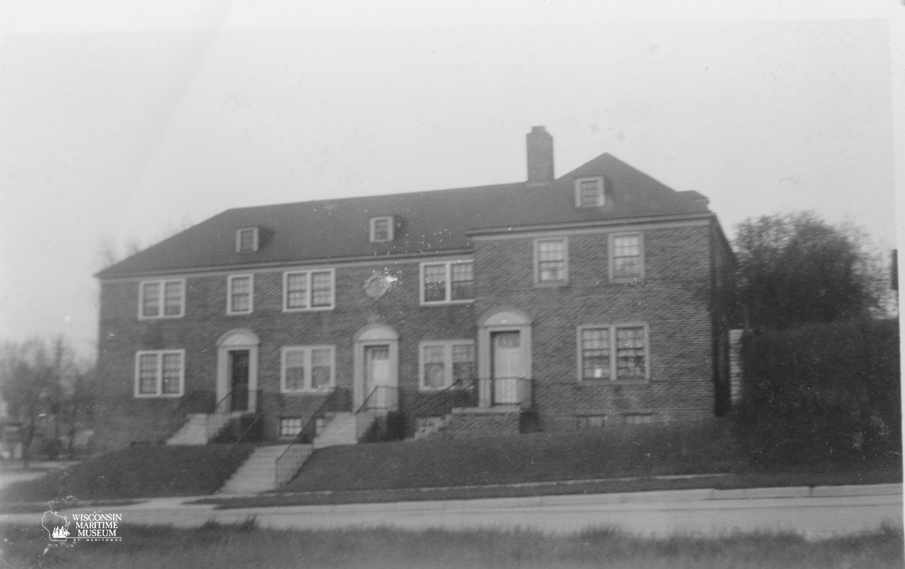 A vintage black-and-white photo of the Manitowoc North Breakwater Lighthouse keeper’s residence circa 1942-1952, a two-story brick building with four front doors, small stairs and a chimney. The setting appears calm and residential.
