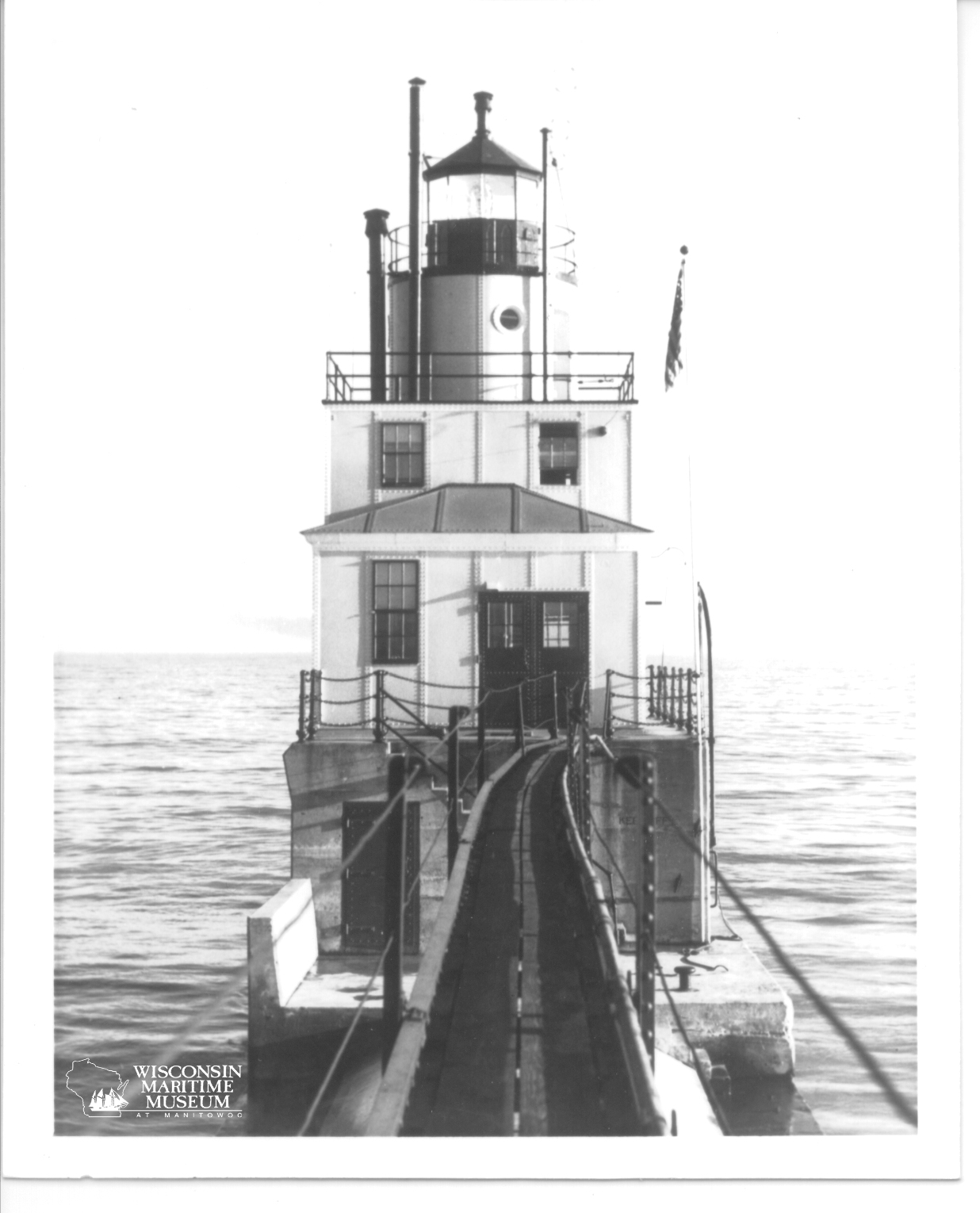 Black and white photo of the Manitowoc North Breakwater Lighthouse circa 1945 with a long walkway jutting from its base into the calm sea. The structure is imposing and symmetrical.