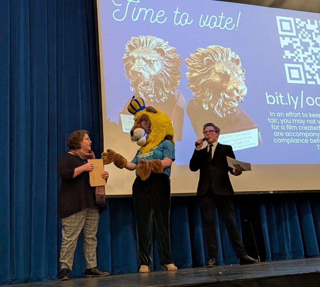 A woman, a lion school. mascot, and a student stand on a stage in front of a projection screen.