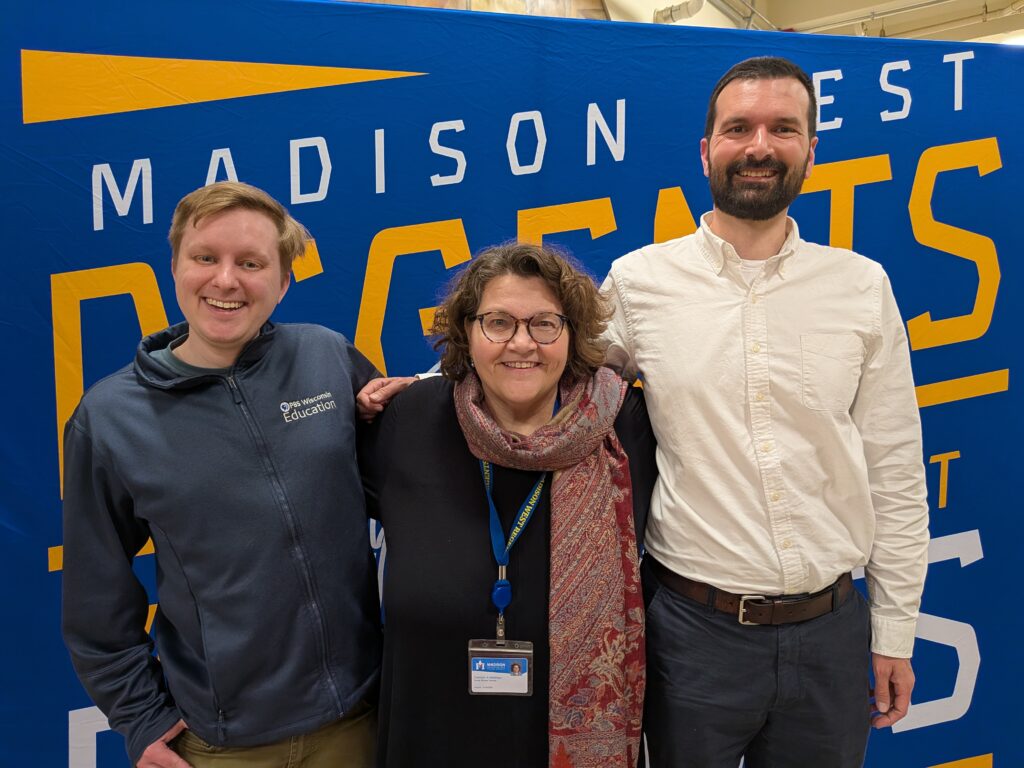 Three people stand with their arms around each other's shoulders, smiling at the camera.
