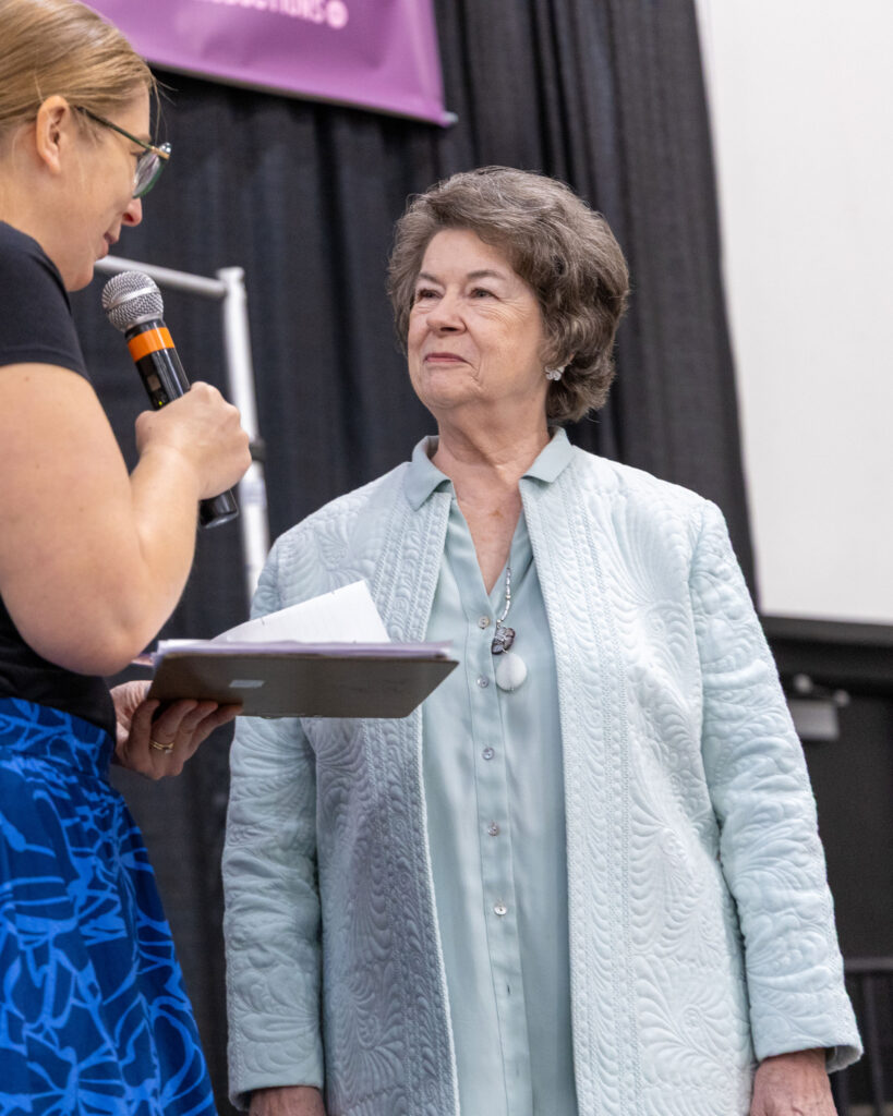 A woman smiles as she is interviewed on stage wearing a light blue, quilted jacket. 