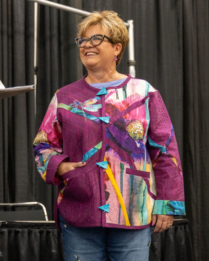 A woman wearing a pink and floral quilted jacket smiles on stage.