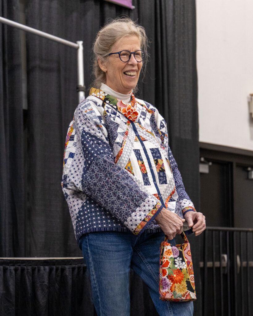 A woman smiles on stage wearing a colorful quilted jacket with a matching floral purse.