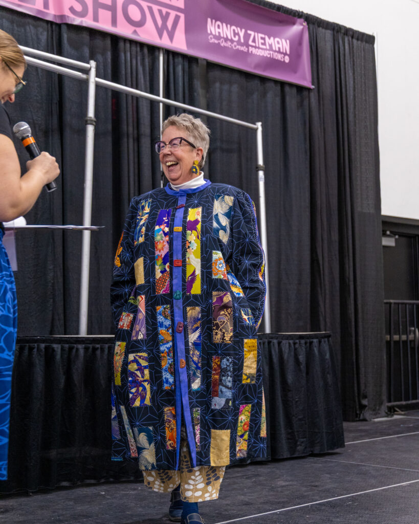 A woman smiles on stage wearing a long, quilted jacket.