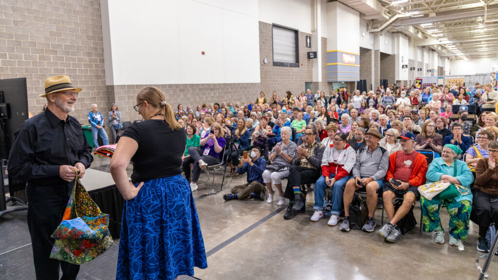 A man is interviewed by a woman on stage as he holds a colorful garment. They are watched by a packed, attentive crowd in a convention center.
