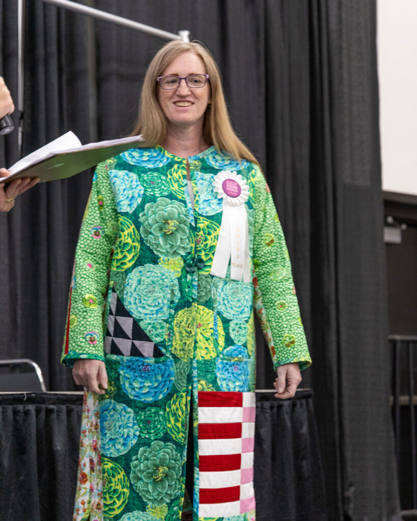 A woman wearing a colorful floral jacket with a white, third-place ribbon stands on stage.