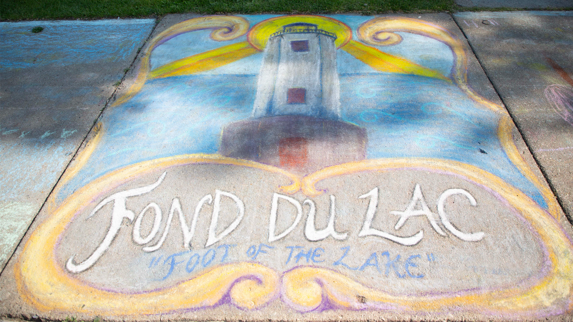 Colorful sidewalk chalk art of a lighthouse with "Fond du Lac" and "Foot of the Lake" written below it, surrounded by swirls and sky elements.