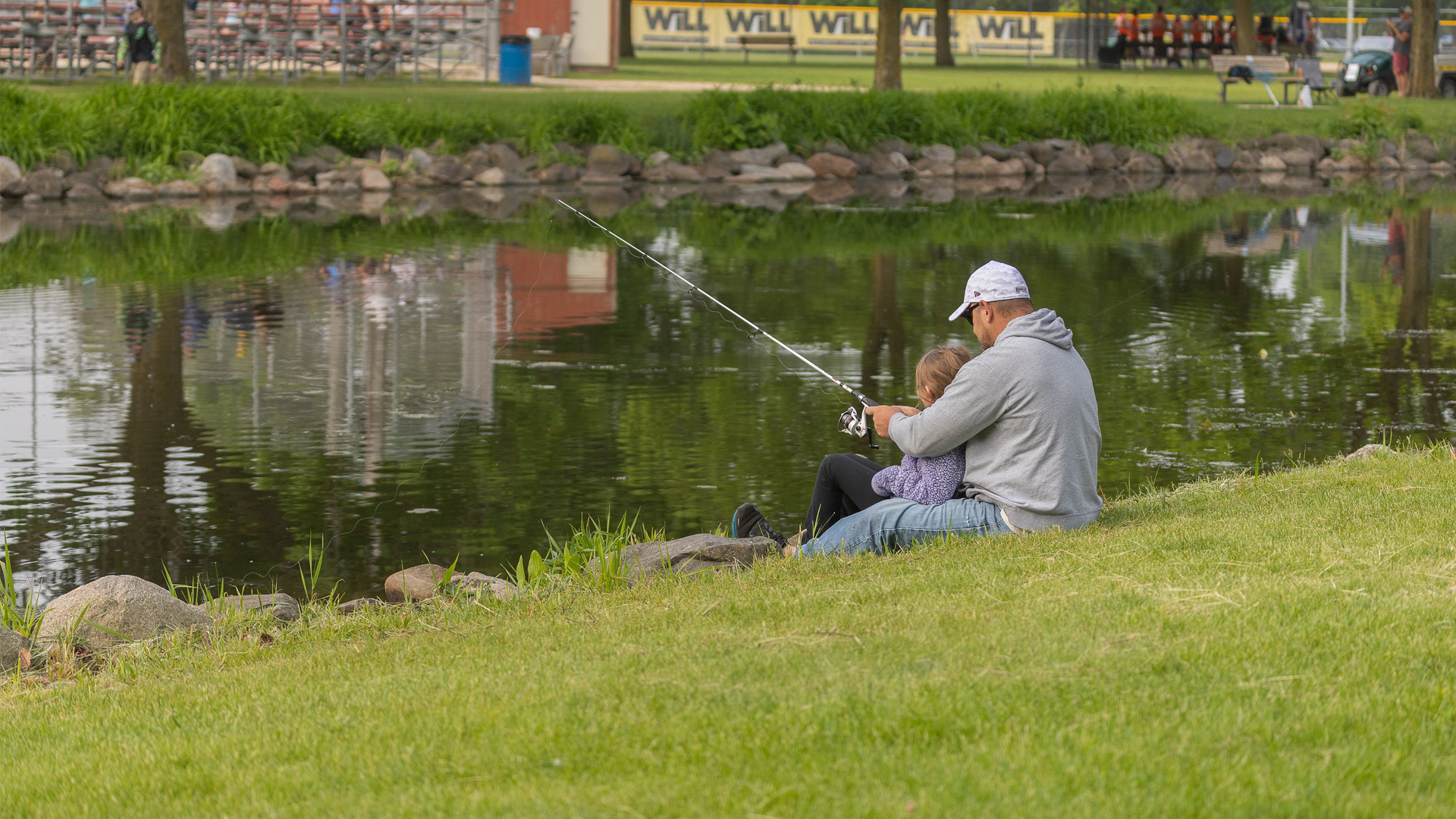 A man and child sit on a grassy bank fishing by a calm pond. The scene is peaceful and reflective, with greenery and distant buildings visible.