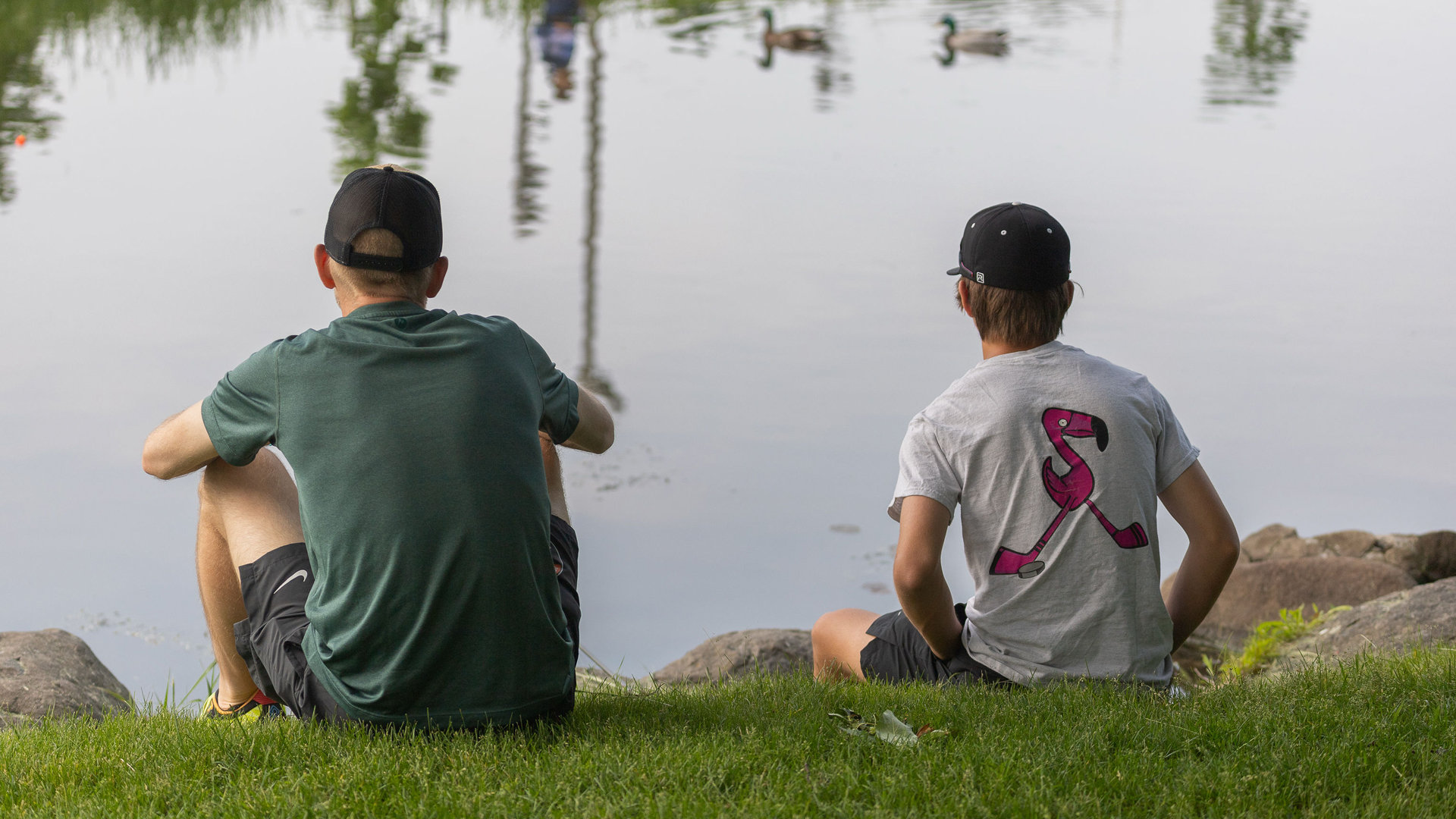 Two people wearing caps sit on grass near a calm lake. Ducks swim in the background, creating a peaceful and relaxing atmosphere.