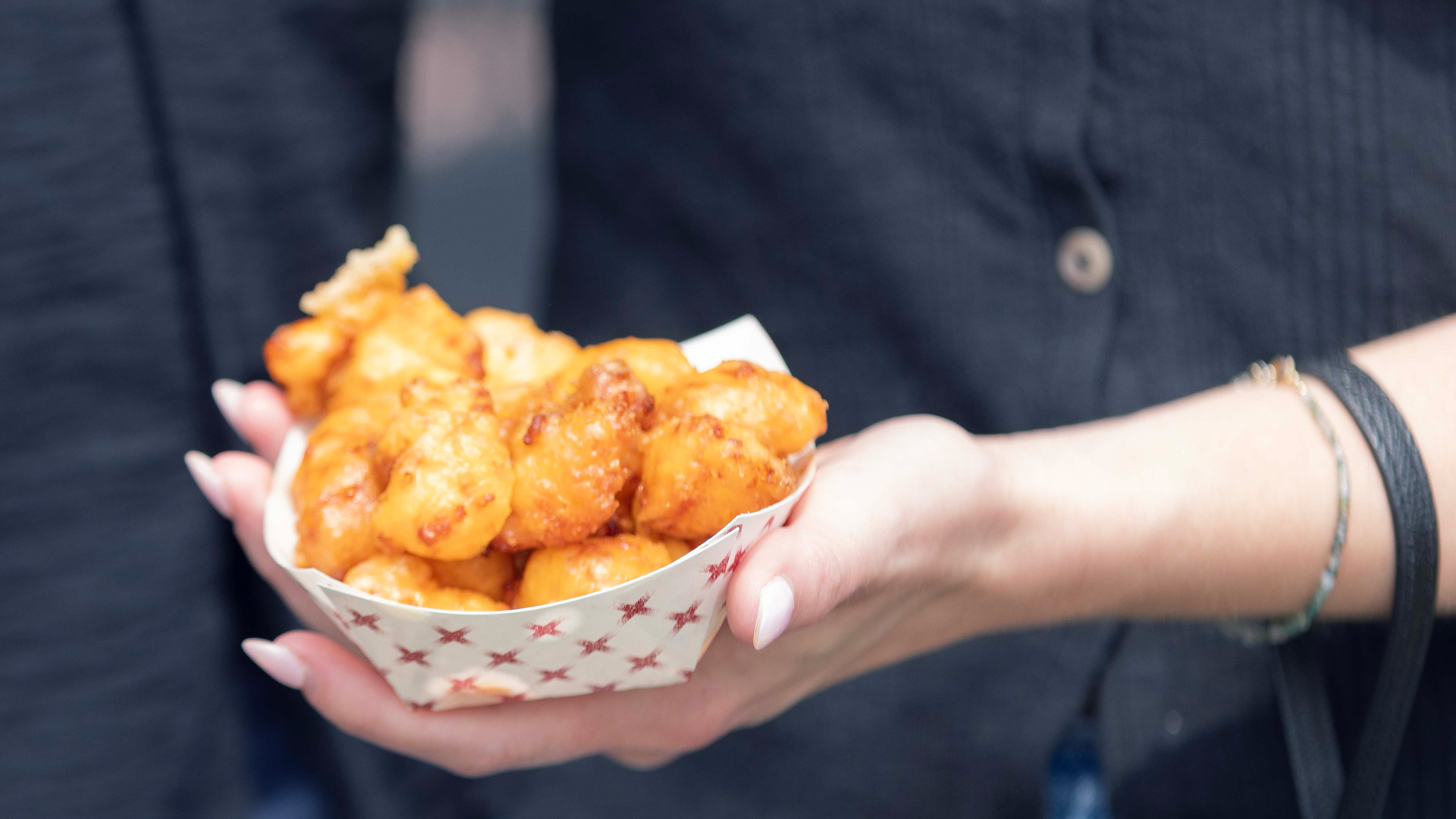 A person holds a tray of golden-brown cheese curds in a paper cup with a red star pattern, against a blurred dark background, conveying a street food vibe.