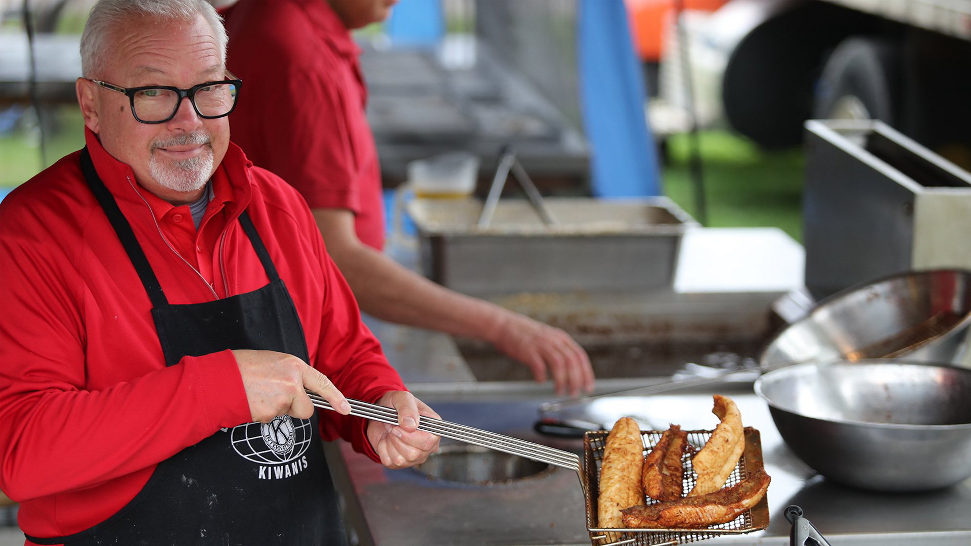 An older man in glasses and a red shirt, wearing a black apron, smiles while holding a basket of fried fish at an outdoor food stand.