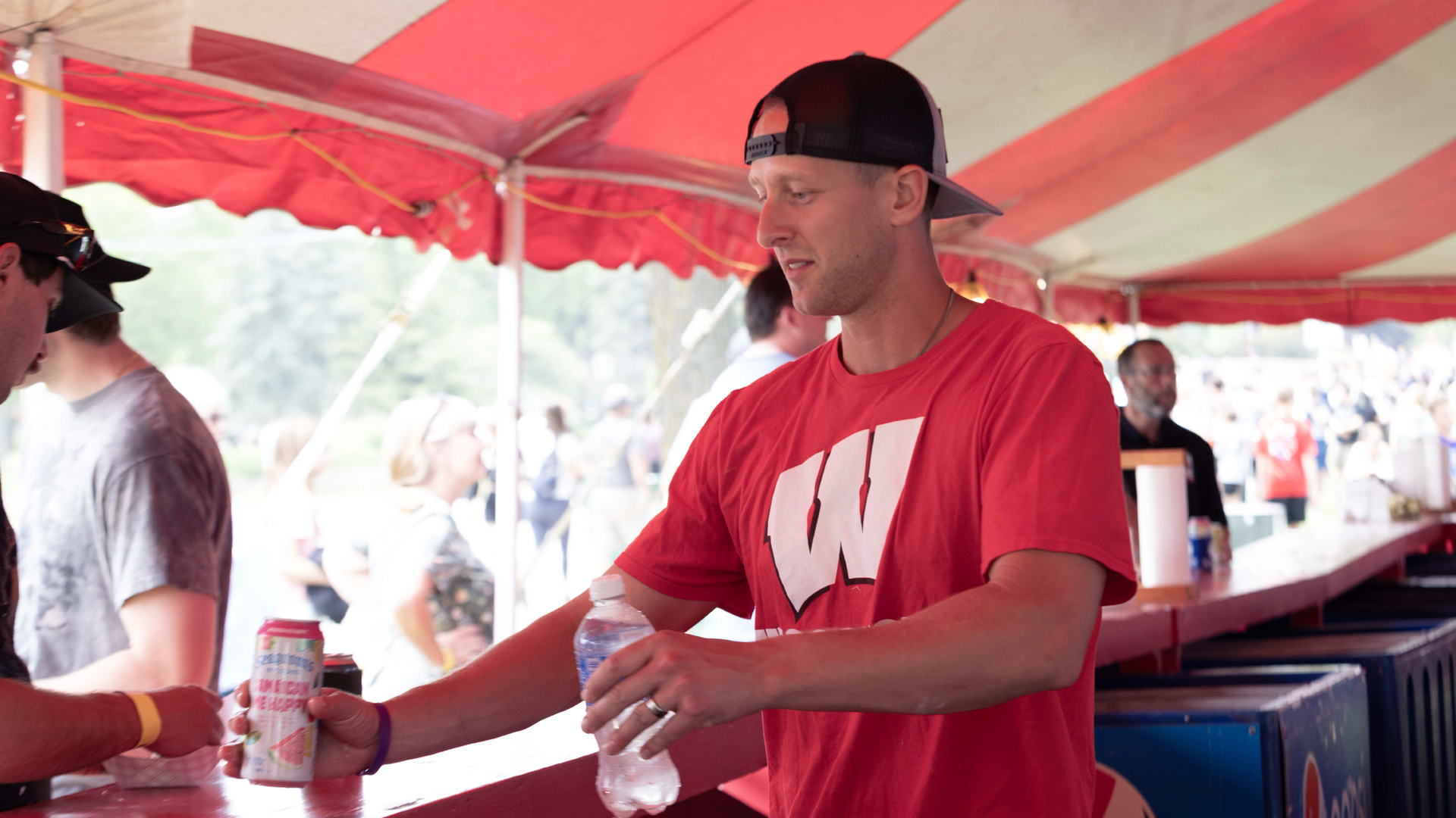 A man in a red t-shirt and backward cap serves drinks at a festival under a red tent. He smiles while handing beverages to a guest, conveying a friendly atmosphere.