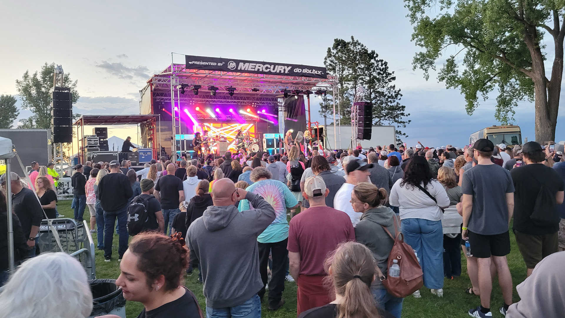 A lively outdoor concert scene with a diverse crowd facing a brightly lit stage. Colorful lights illuminate performers, and a relaxed, festive atmosphere prevails.