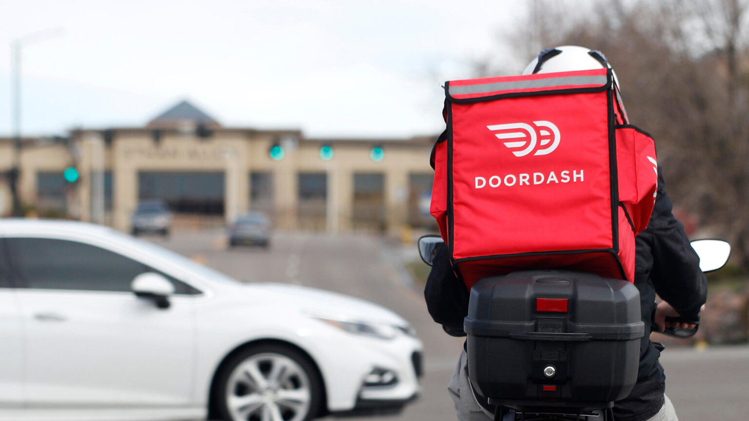 A person driving a moped with a tailbox and wearing an insulated square backpack with side pockets and the DoorDash logo and word DOORDASH faces an intersection with an out-of-focus sedan passing through it, with an out-of-focus building, traffic lights, driving vehicles and leafless trees in the background.