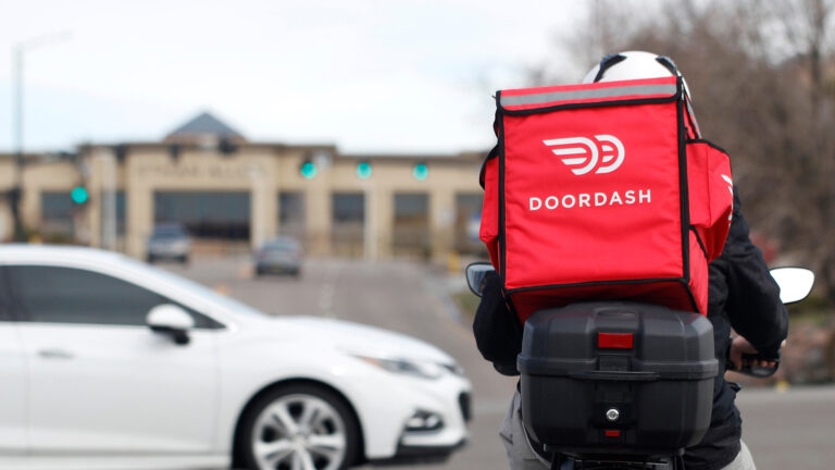 A person driving a moped with a tailbox and wearing an insulated square backpack with side pockets and the DoorDash logo and word DOORDASH faces an intersection with an out-of-focus sedan passing through it, with an out-of-focus building, traffic lights, driving vehicles and leafless trees in the background.