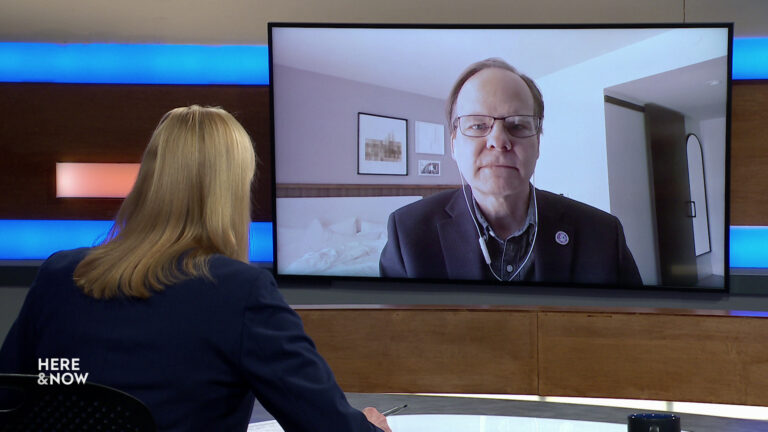 Frederica Freyberg sits at a desk on the Here & Now set and faces a video monitor showing an image of Tom Content.