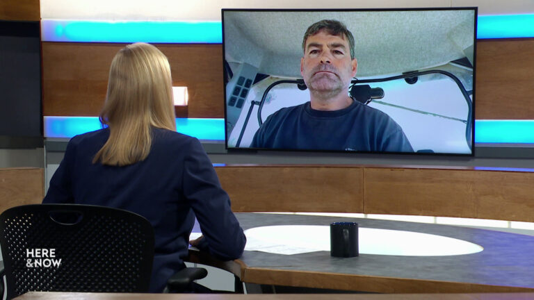 Frederica Freyberg sits at a desk on the Here & Now set and faces a video monitor showing an image of Tony Kurtz.