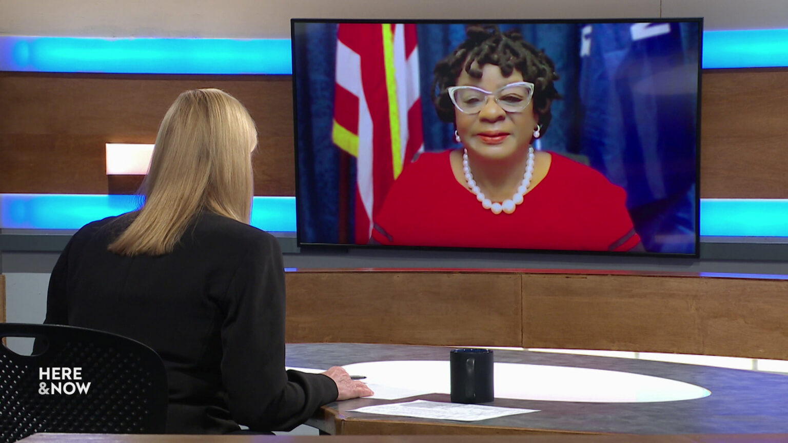 Frederica Freyberg sits at a desk on the Here & Now set and faces a video monitor showing an image of Gwen Moore.
