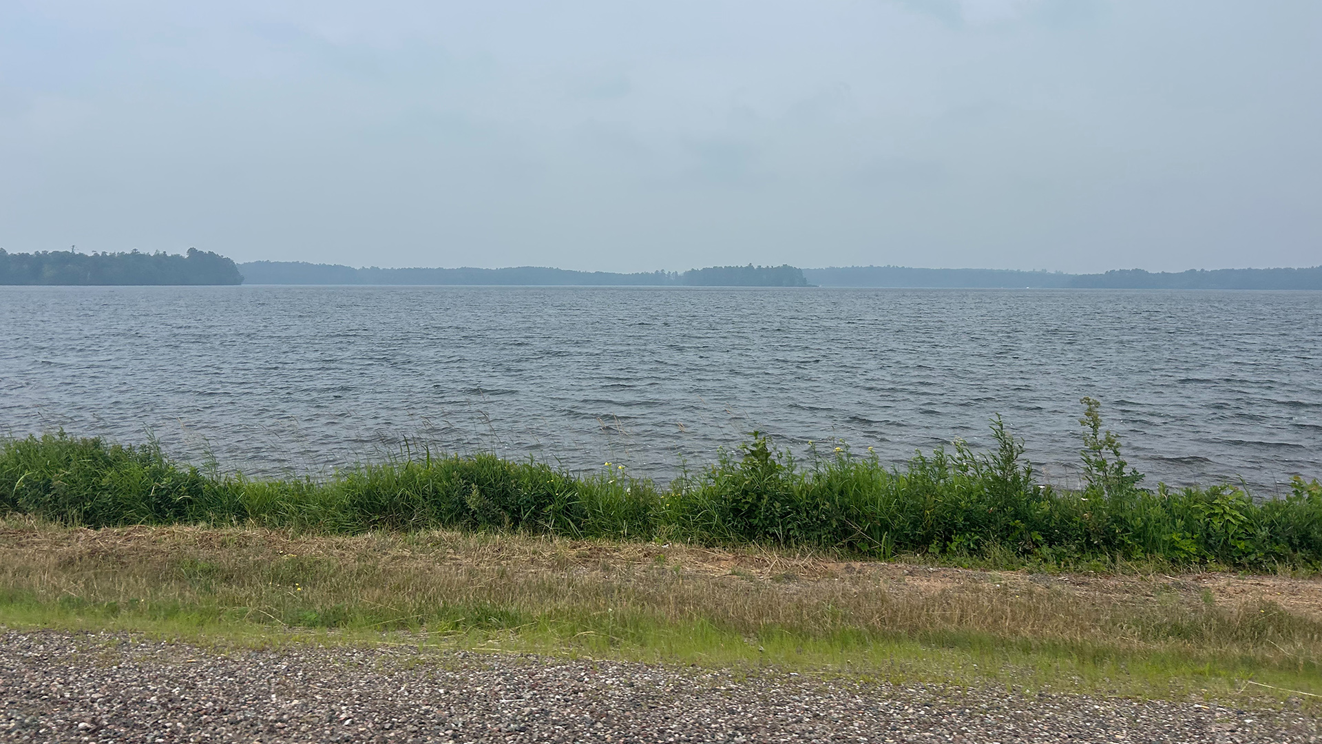 Small waves on a lake move toward its near shore, with plants, a mowed area and a gravel track in the foreground, and the hazy tree-covered far shore on the horizon in the background.