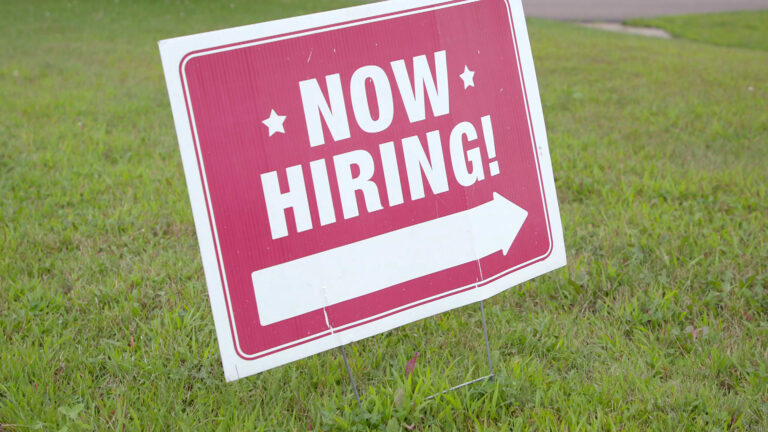 A cardboard sign with the text Now Hiring! and an arrow pointing to the right stands in a lawn.