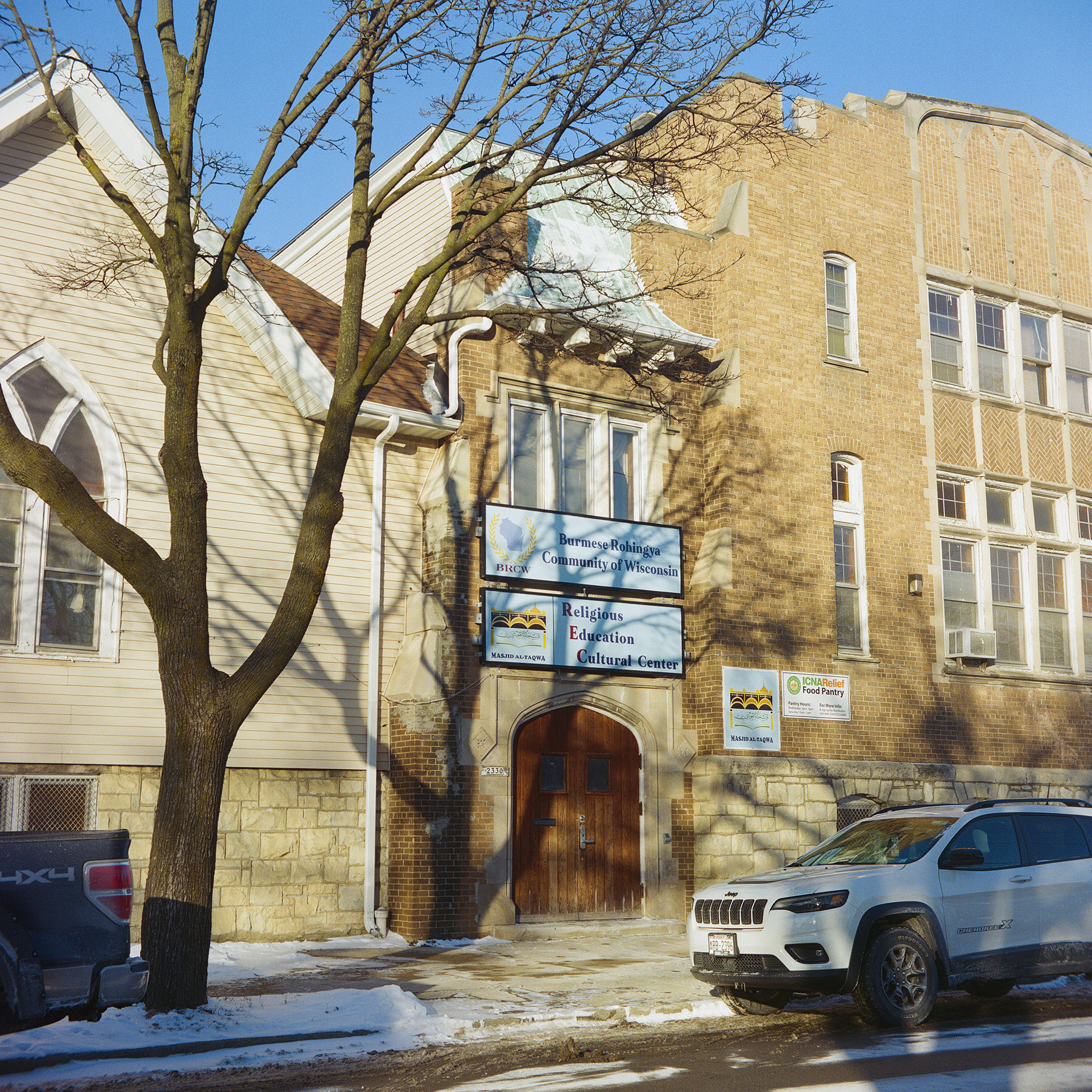 Two vehicles are parked next to a curb with a leafless tree in a snowbank on one side in front of a multi-wing and multi-story masonry and brick building with a double wood door, a gothic arch window on one side and rectangular windows on the other side, and signs reading "Burmese Rohingya Community of Wisconsin" and "Religious Education Cultural Center."