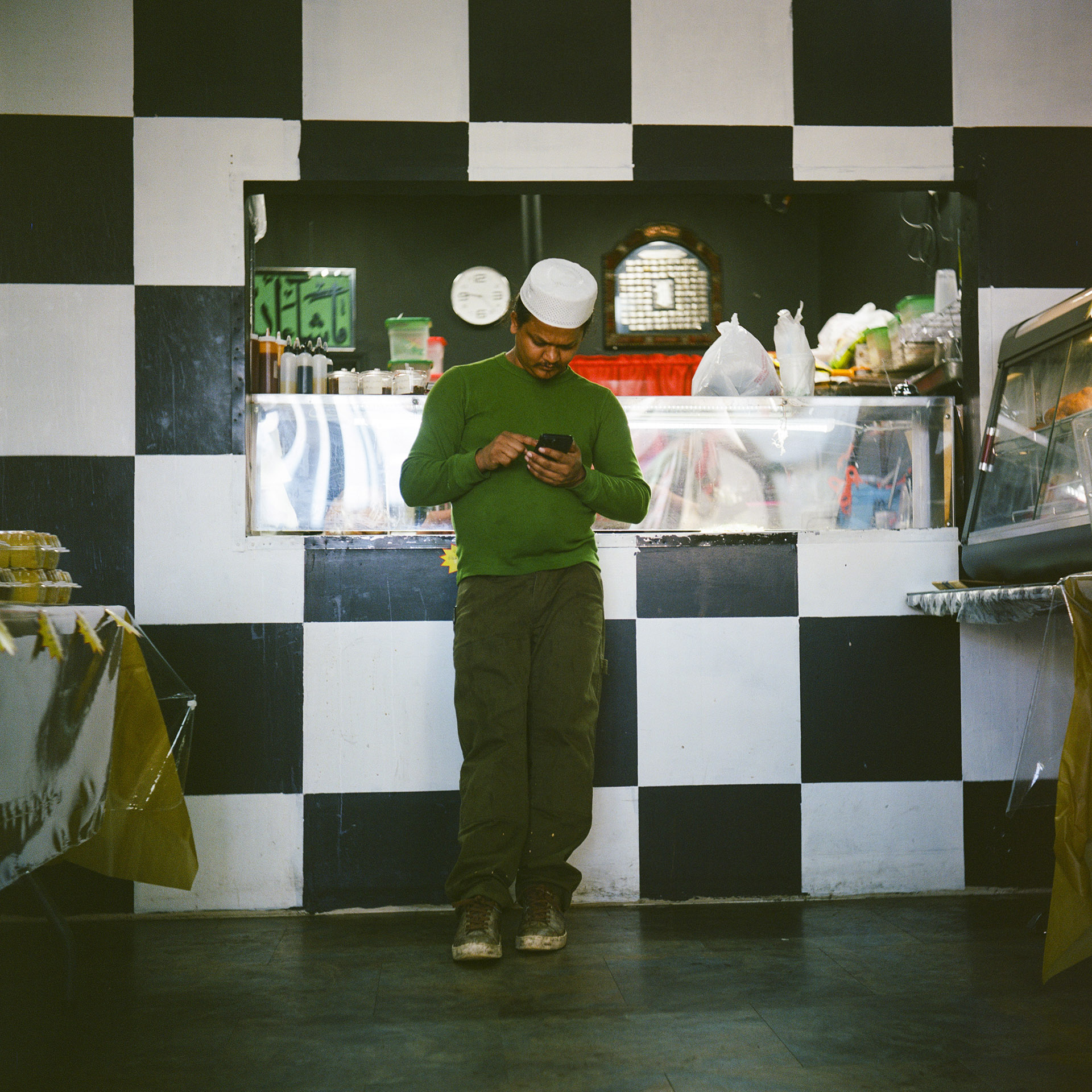 Mohamed Ibrahim uses a smart phone in his hands while leaning back against a checkerboard-painted wall with a serving window with a display case at its bottom and other items visible above it on the rear wall, and with a table and another food display case on either side.