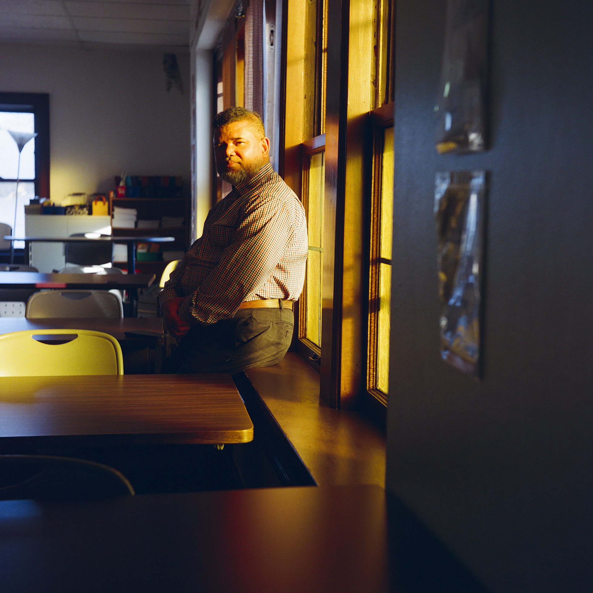 Mohamed Anwar poses for a portrait while sitting on a windowsill between two desks, with light from a row of open windows illuminating his back, with a table, lamp and another window on a far wall in the background.