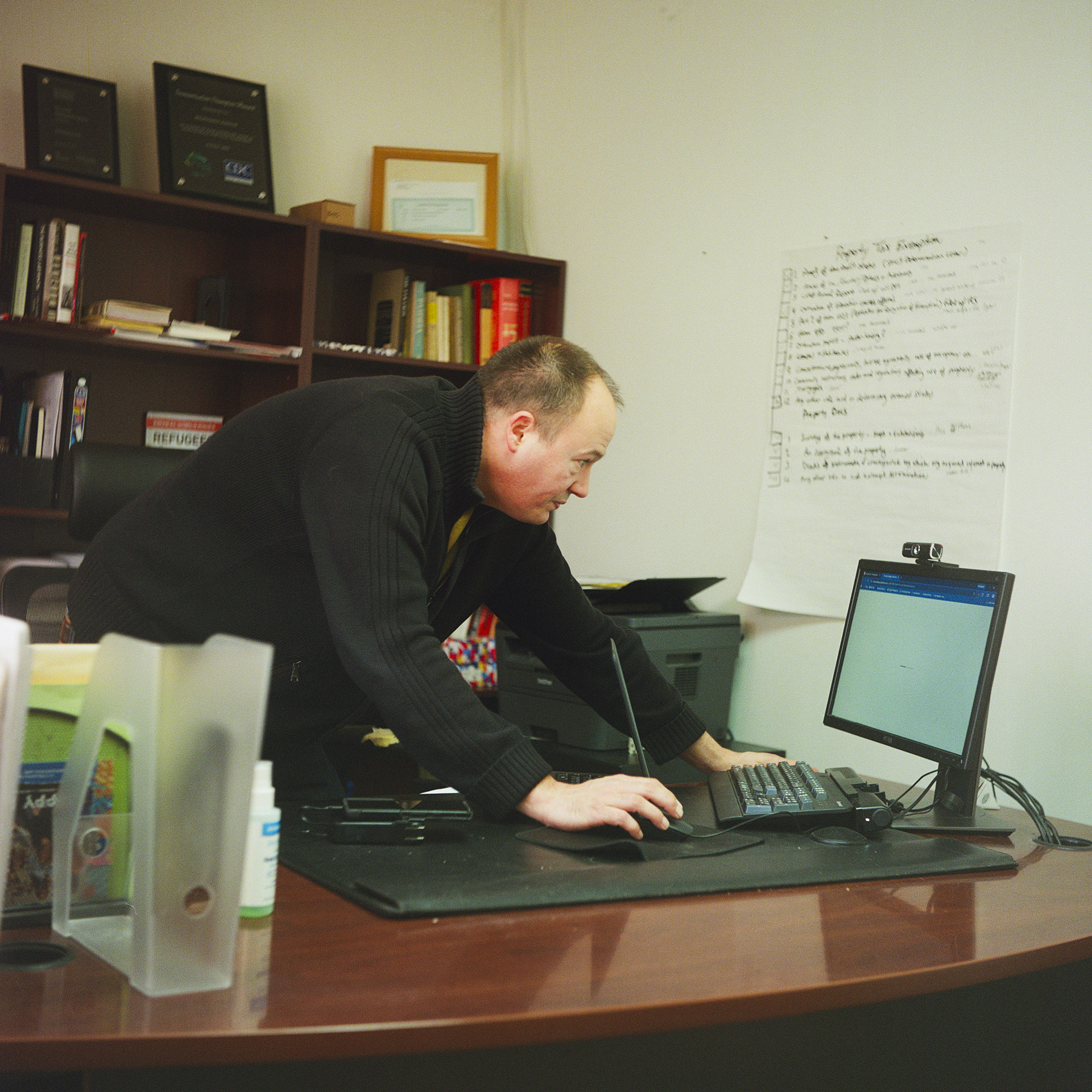 Andrew Trumbull stands and leans over a desk while using moving a wired computer mouse with his right hand and facing a keyboard and computer monitor on a wood desk, with bookshelves with books and plaques behind him and a large piece of paper with writing on it mounted on a wall.