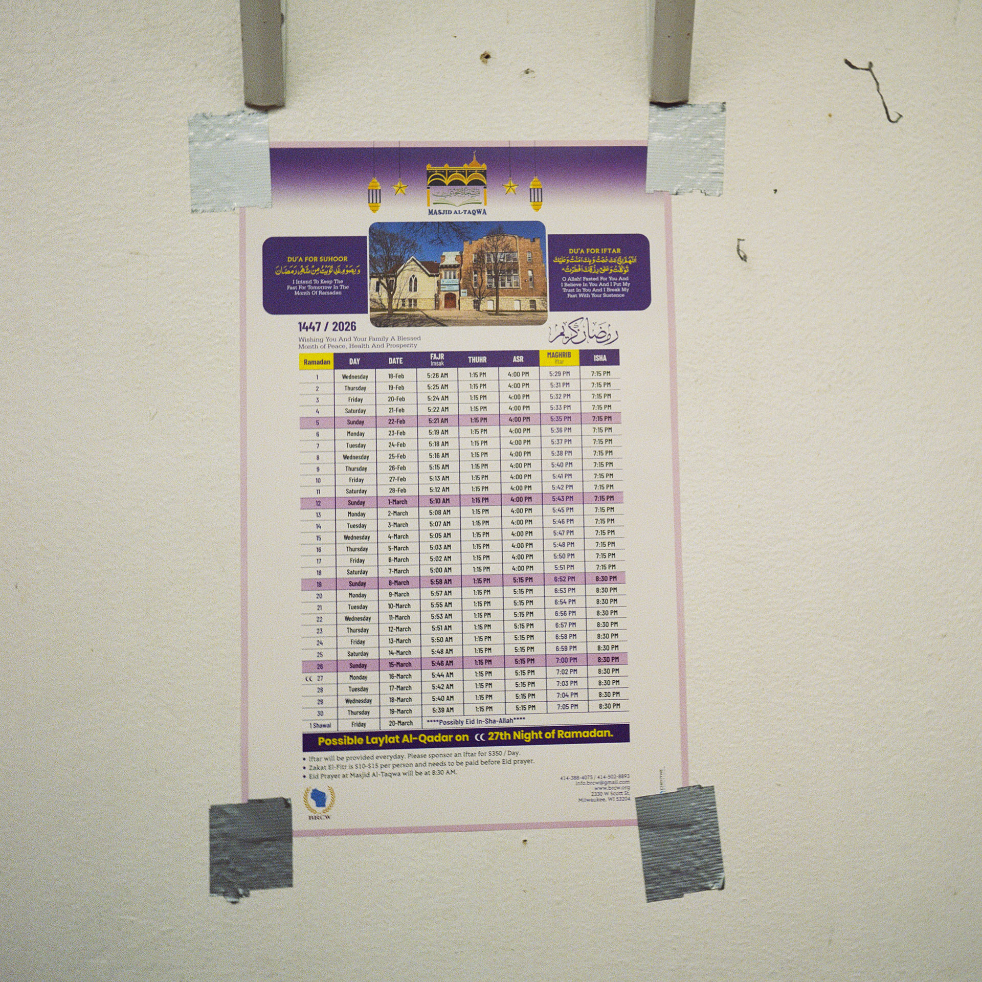 A calendar with a timetable is mounted to a wall with four pieces of duct tape.