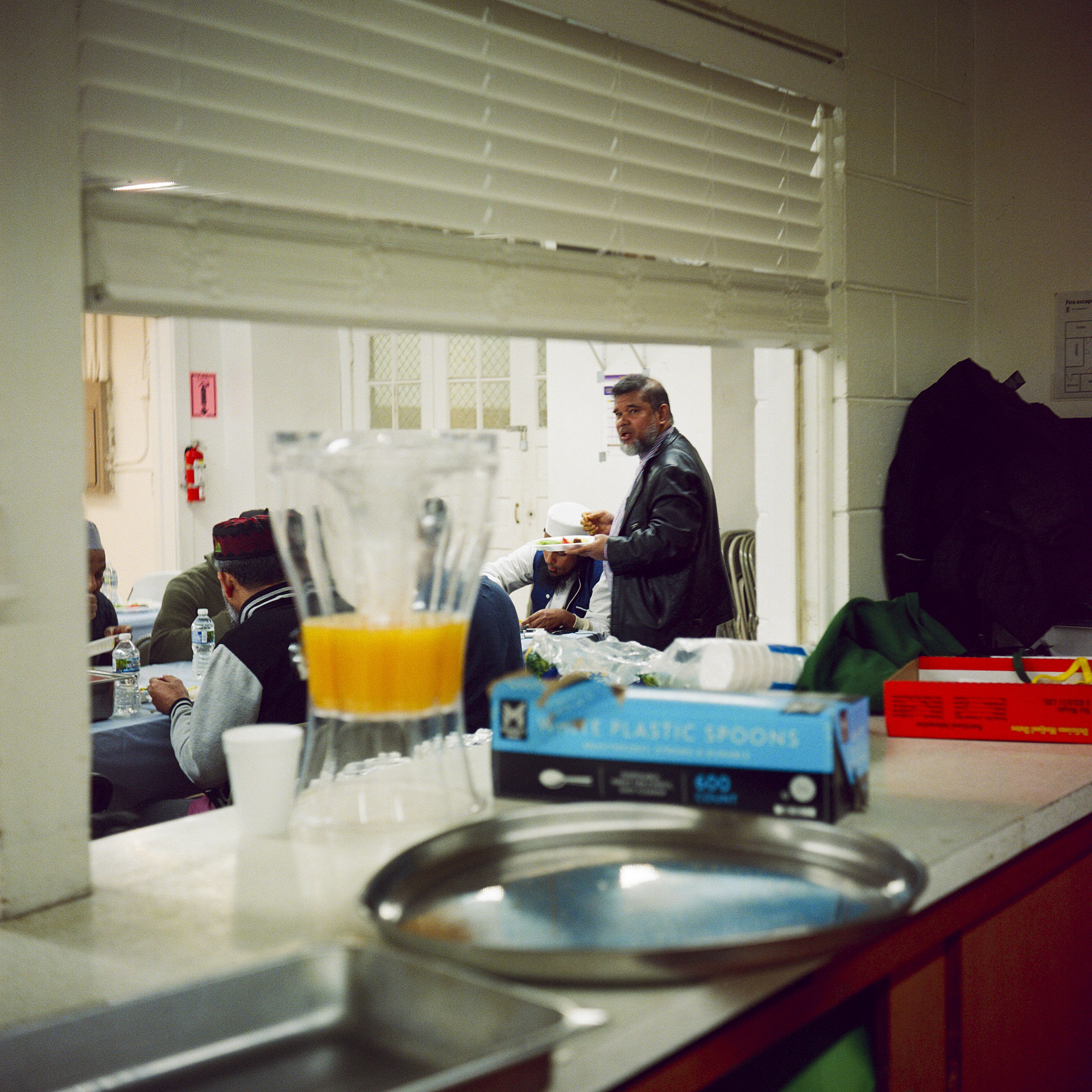Mohamed Anwar holds a plate of food in his left hand while standing next to a table with men seated around it, as ween through a serving window of a kitchen with raised blinds and an out-of-focus beverage dispenser, metal serving tray and other items on a countertop in the foreground.