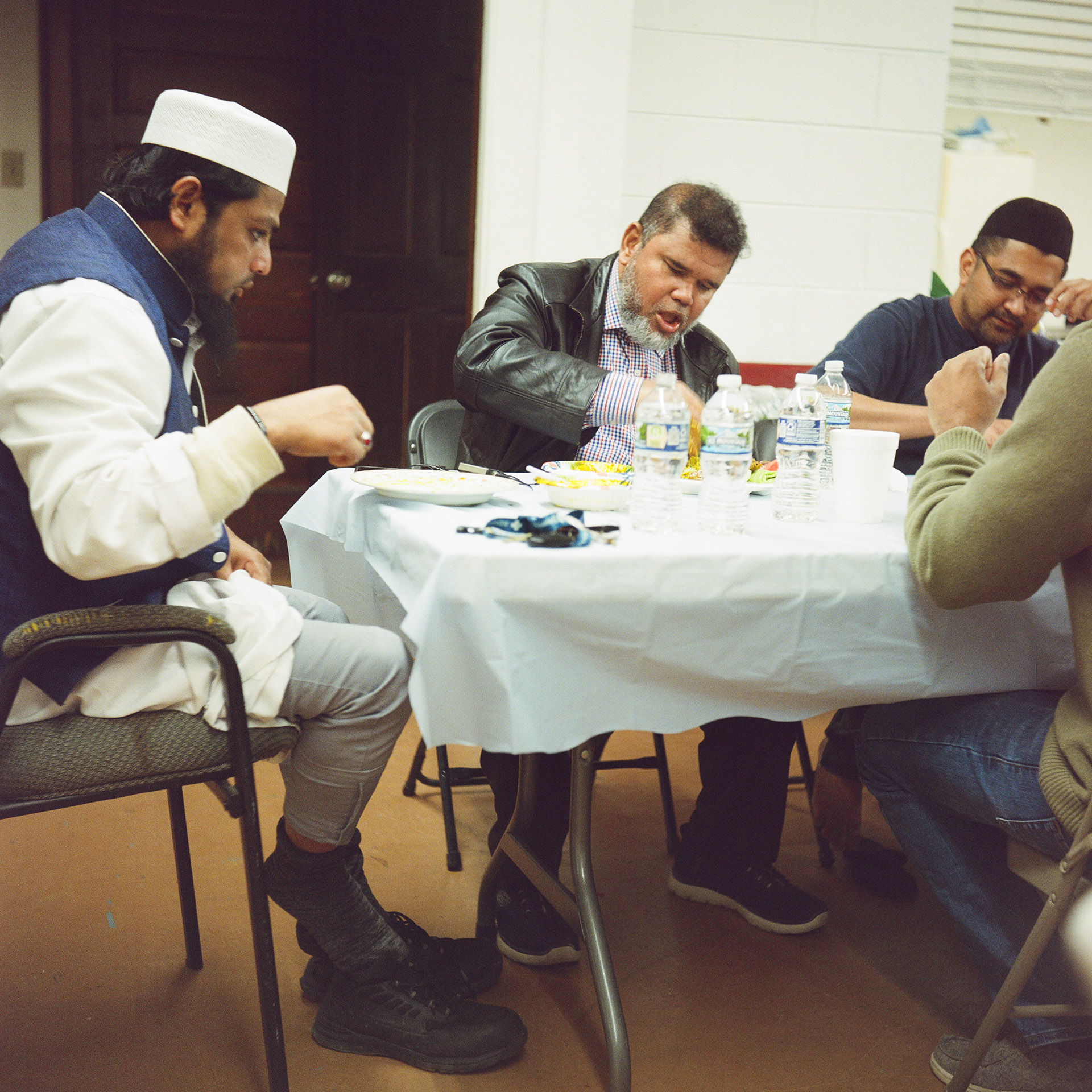Mohamed Anwar sits at a folding table covered with a tablecloth and multiple plastic water bottles on its surface, with other men seated on either side and across from him, in a room with a closed wood door and painted concrete block walls in the background.