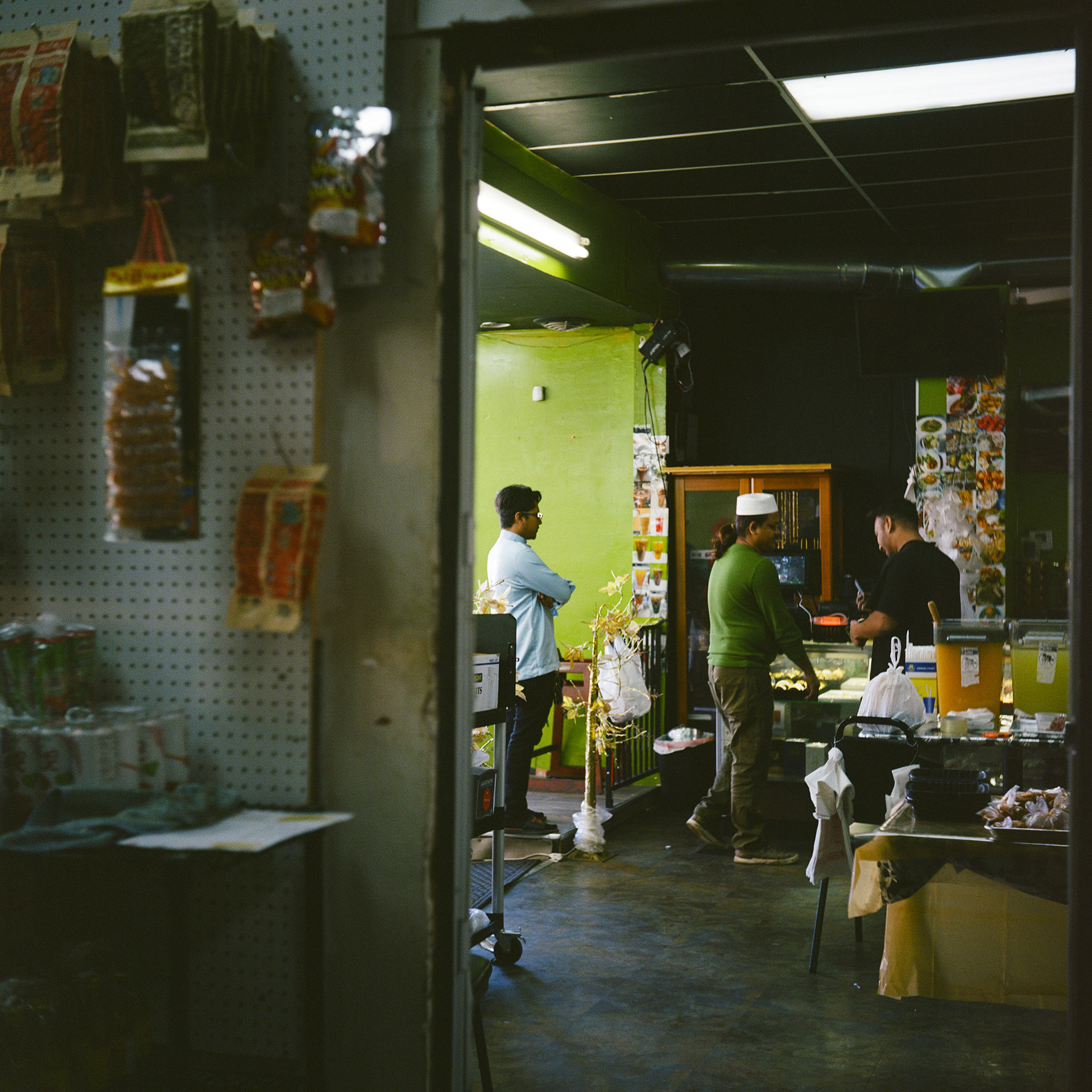 Mohamed Ibrahim stands in front of a food display case and talks to another person, with a third person standing behind them, in a room with two beverage dispensers on a table, a rolling table, a wood-and-glass cabinet and other items, with fluorescent lights in a drop-panel ceiling, as ween through the threshold of a doorway with out-of-focus food items displayed on a pegboard wall in the foreground.