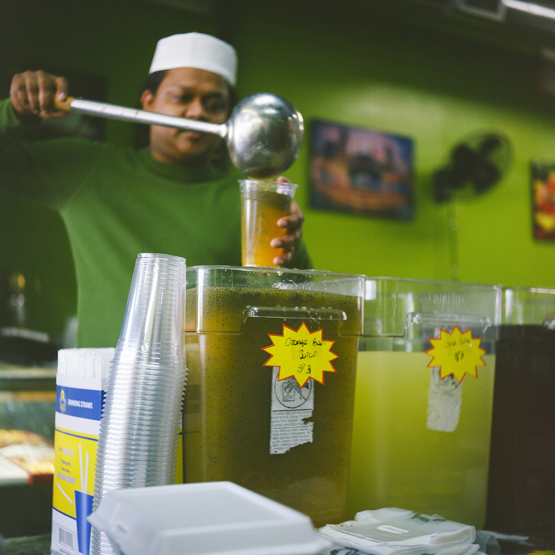 Mohamed Ibrahim uses a metal ladle to dispense a beverage from a plastic container into a disposable plastic cup, with other beverage containers and stack of cups in front of him, in a room with an out-of-focus a wall-mounted fan in the background.